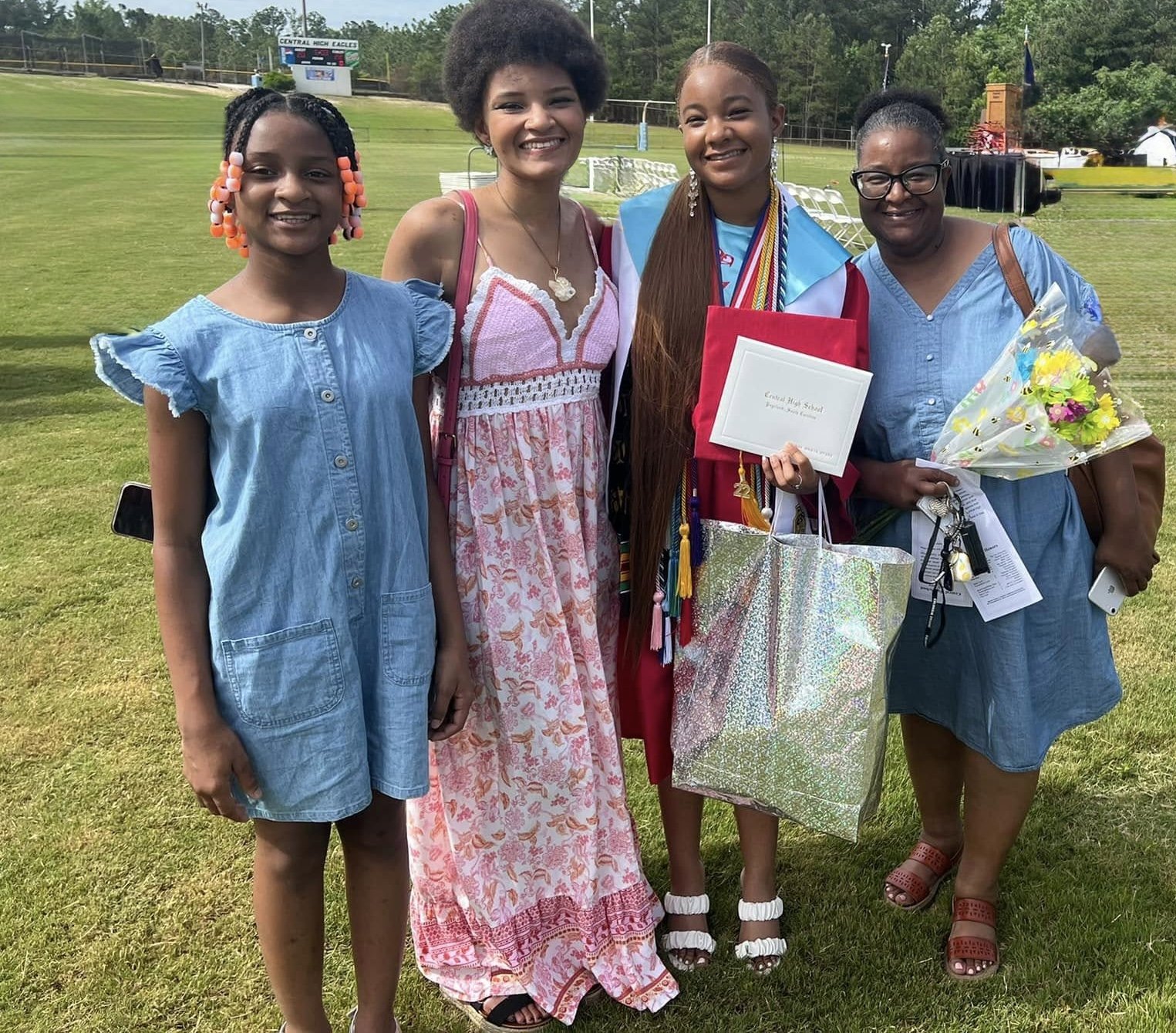 A group of four women and girls standing on a grassy field after a graduation ceremony. The woman in the center is wearing a maroon graduation gown and cap with medals and holding a diploma. The others are dressed in casual summer clothes, smiling at the camera, with one holding a bouquet of flowers.