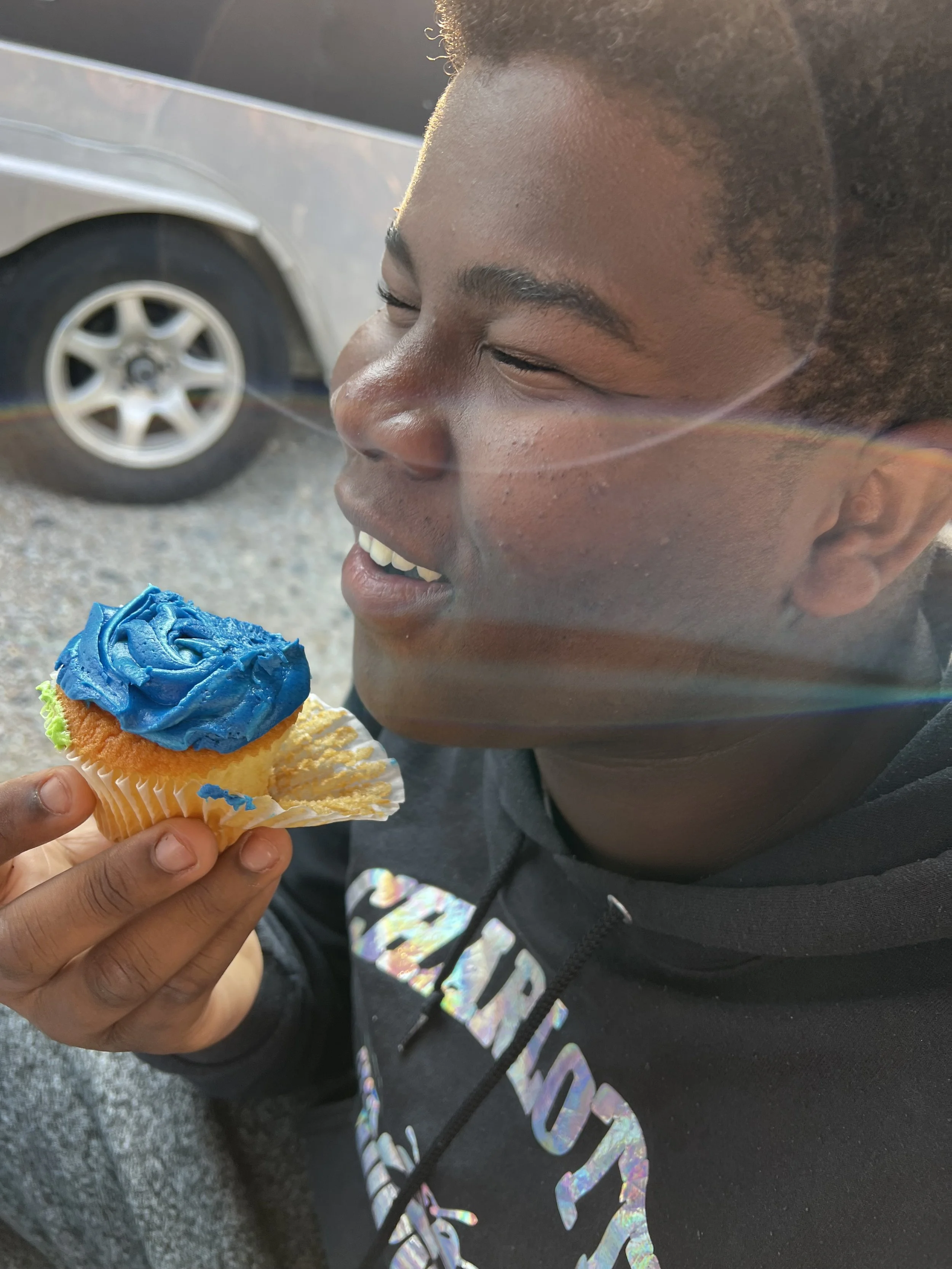 Close-up of a young man with dark skin and a hairstyle with short sides and curly top, smiling with eyes closed, about to bite into a cupcake with blue frosting and sprinkles, held by a hand. In the background, a parked car is visible.