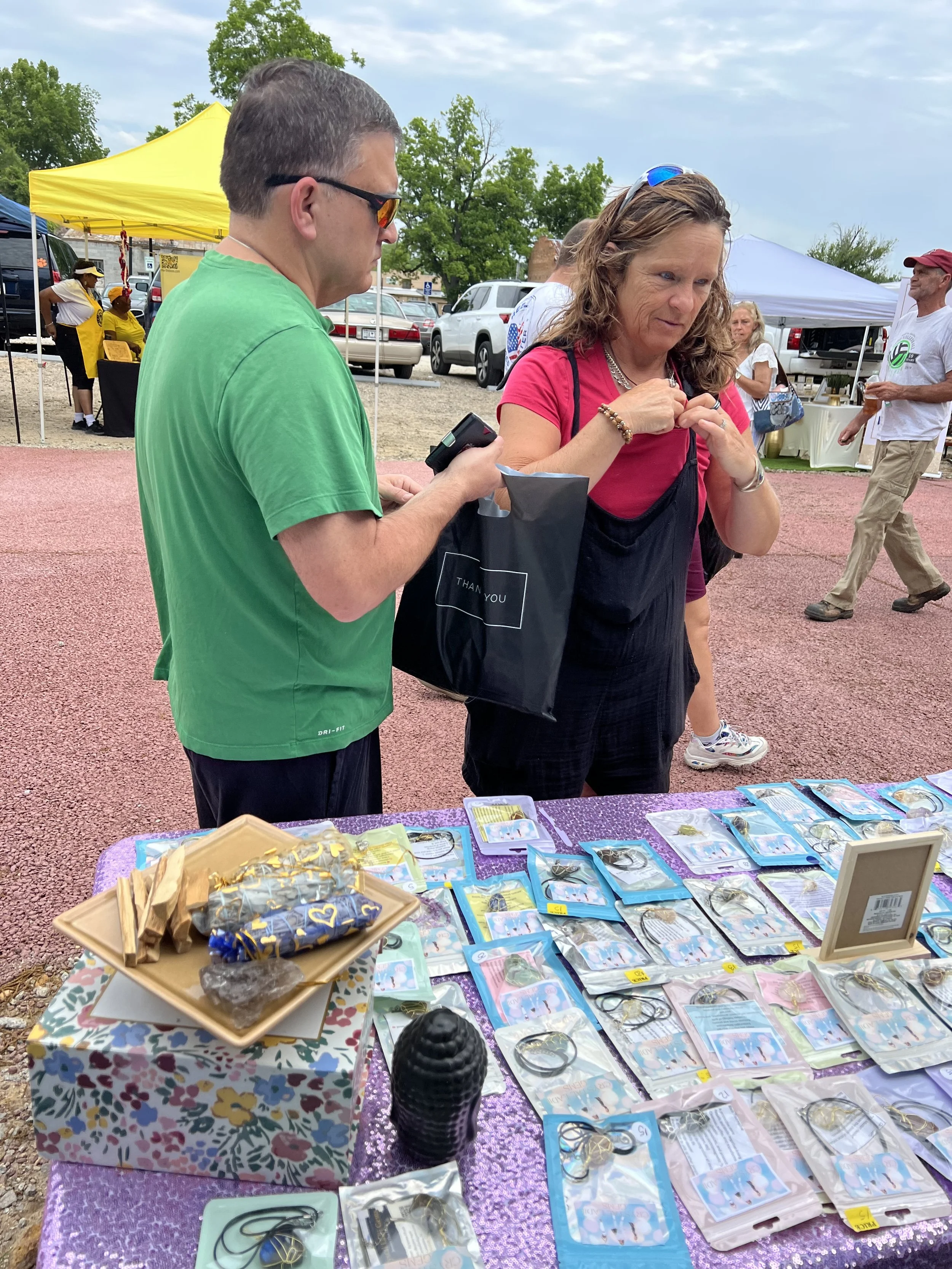 A woman and a man are standing at a table with various jewelry items and accessories for sale at an outdoor market or event. The woman is wearing a pink shirt and an apron, and the man is wearing glasses and a green shirt. The background includes ten