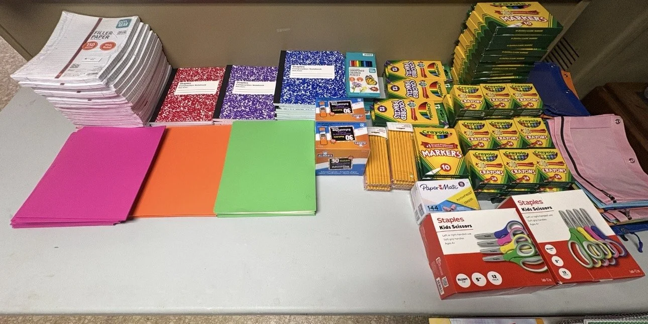 School supplies including notebooks, colored paper, markers, glue sticks, crayons, scissors, and paper.