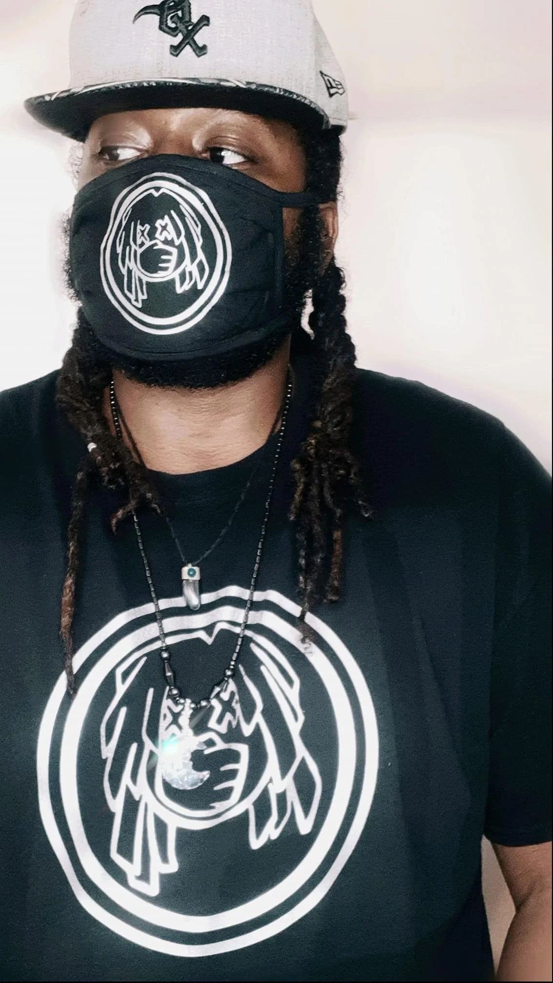 A person wearing a black t-shirt with a white circular graphic of a face with long hair and crossed-out eyes, a matching face mask with the same graphic, a silver necklace with a pendant, and a black and gray cap with a black logo.