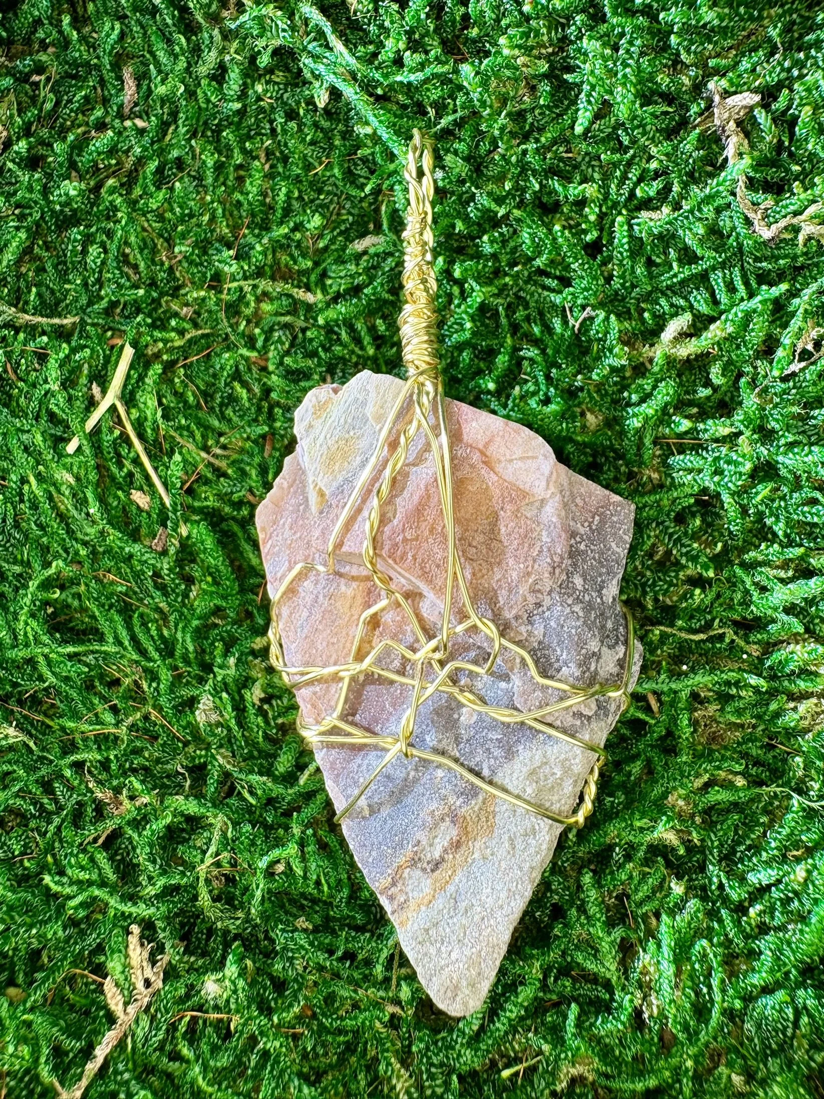 A piece of rough, unpolished pinkish-brown stone on green moss, wrapped in a shiny gold wire.