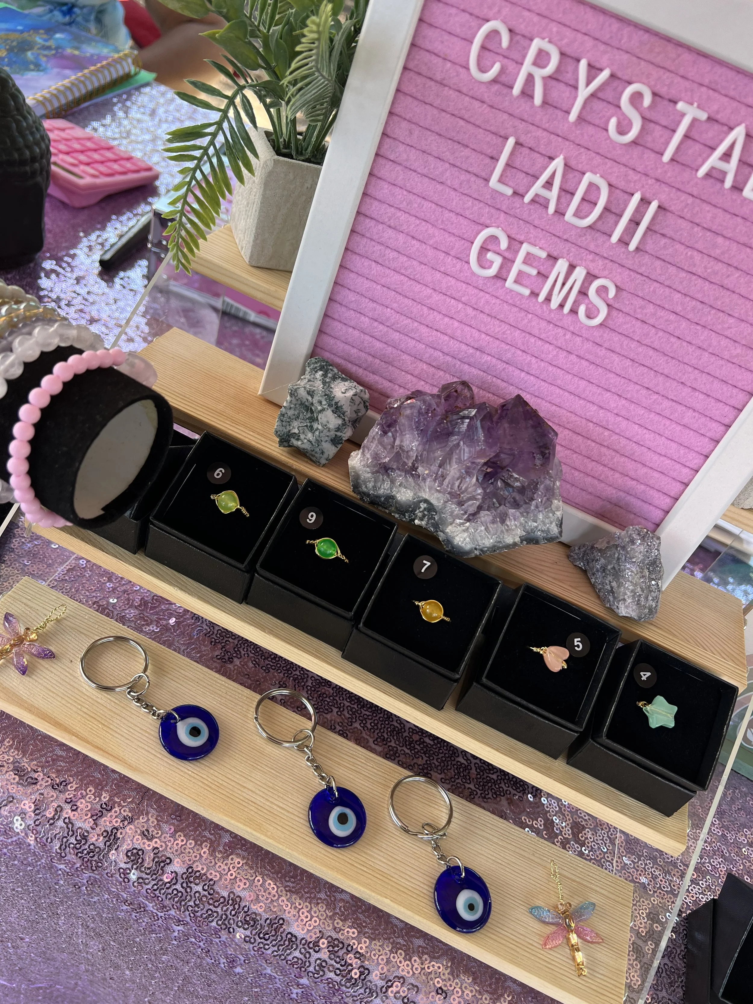 Display of jewelry including rings with colorful stones, earrings shaped like eyes and a dragonfly, along with rocks, crystals, and a pink felt board with captions about crystals, lapis, and gems, on a glittery pink tablecloth.