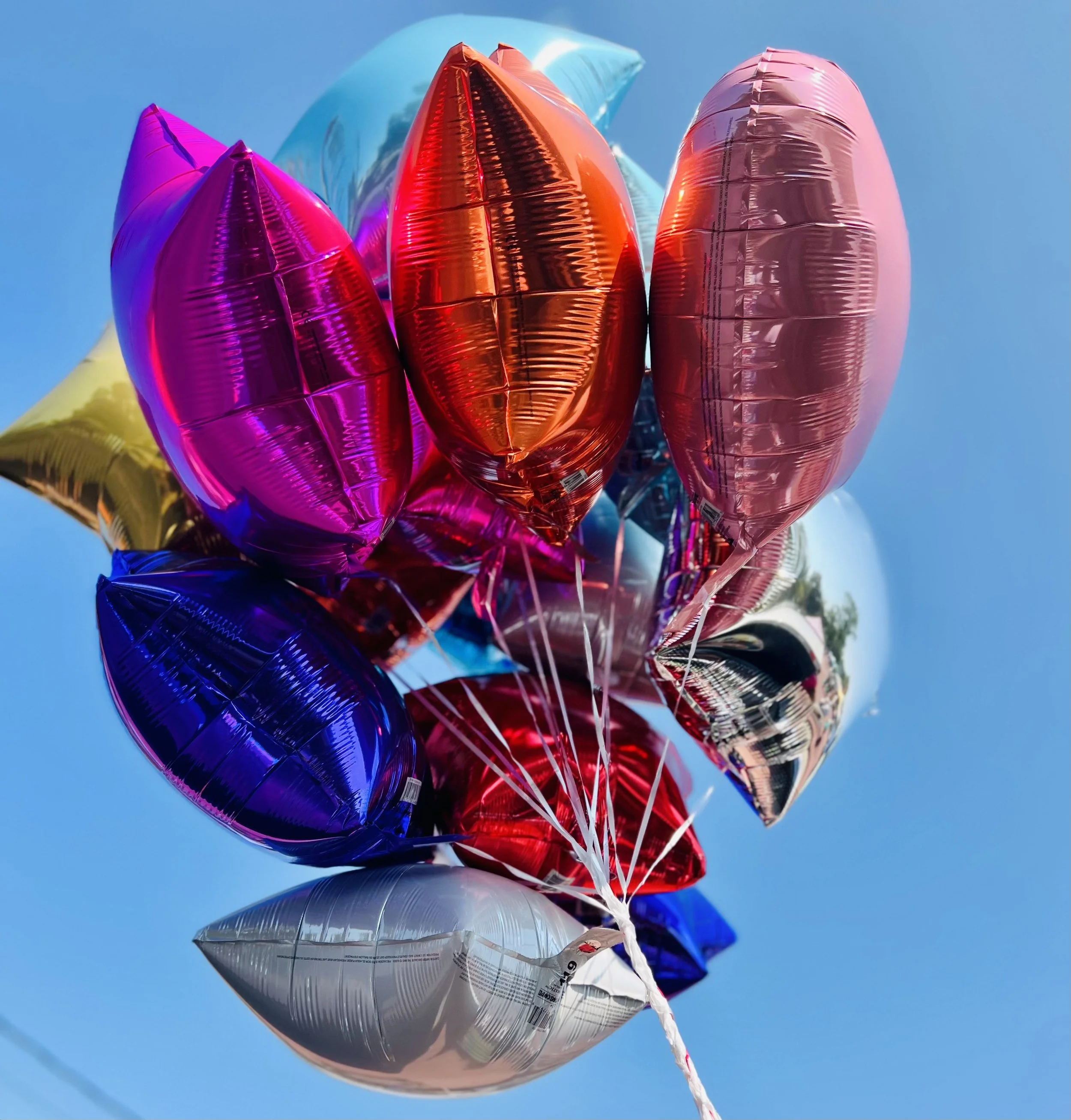 Colorful metallic helium balloons in pink, orange, silver, purple, blue, and yellow, floating against a bright blue sky.