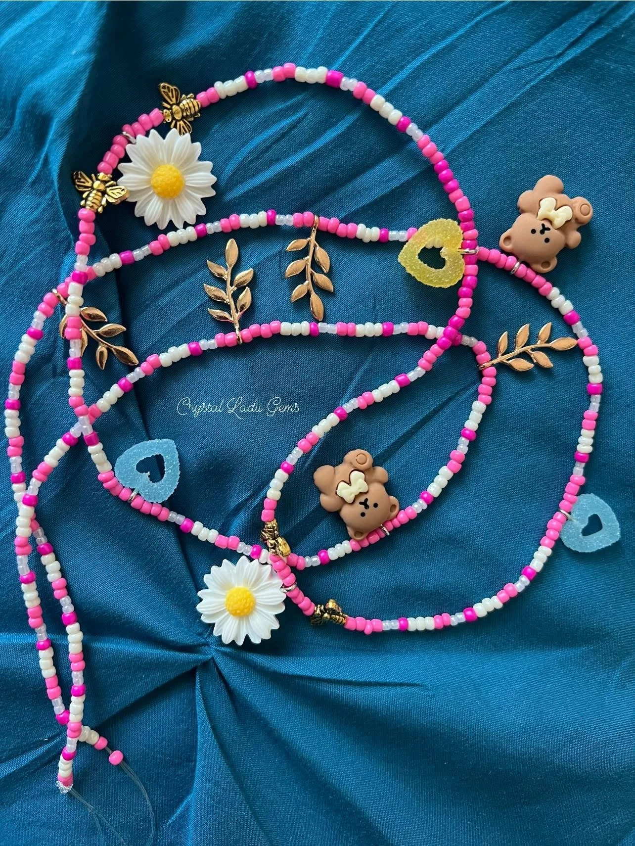 Colorful beaded jewelry necklaces with daisy and teddy bear charms, laid on blue satin fabric with ruffled texture.