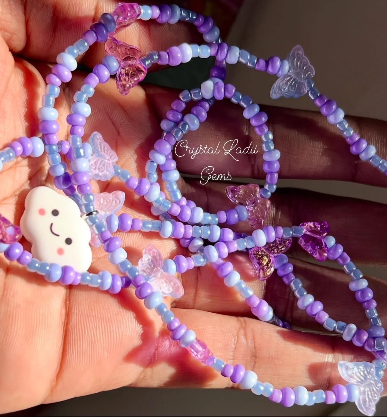 Colorful beaded necklaces with purple, pink, and white beads, featuring butterfly and cloud-shaped charms, held in a person's hand.