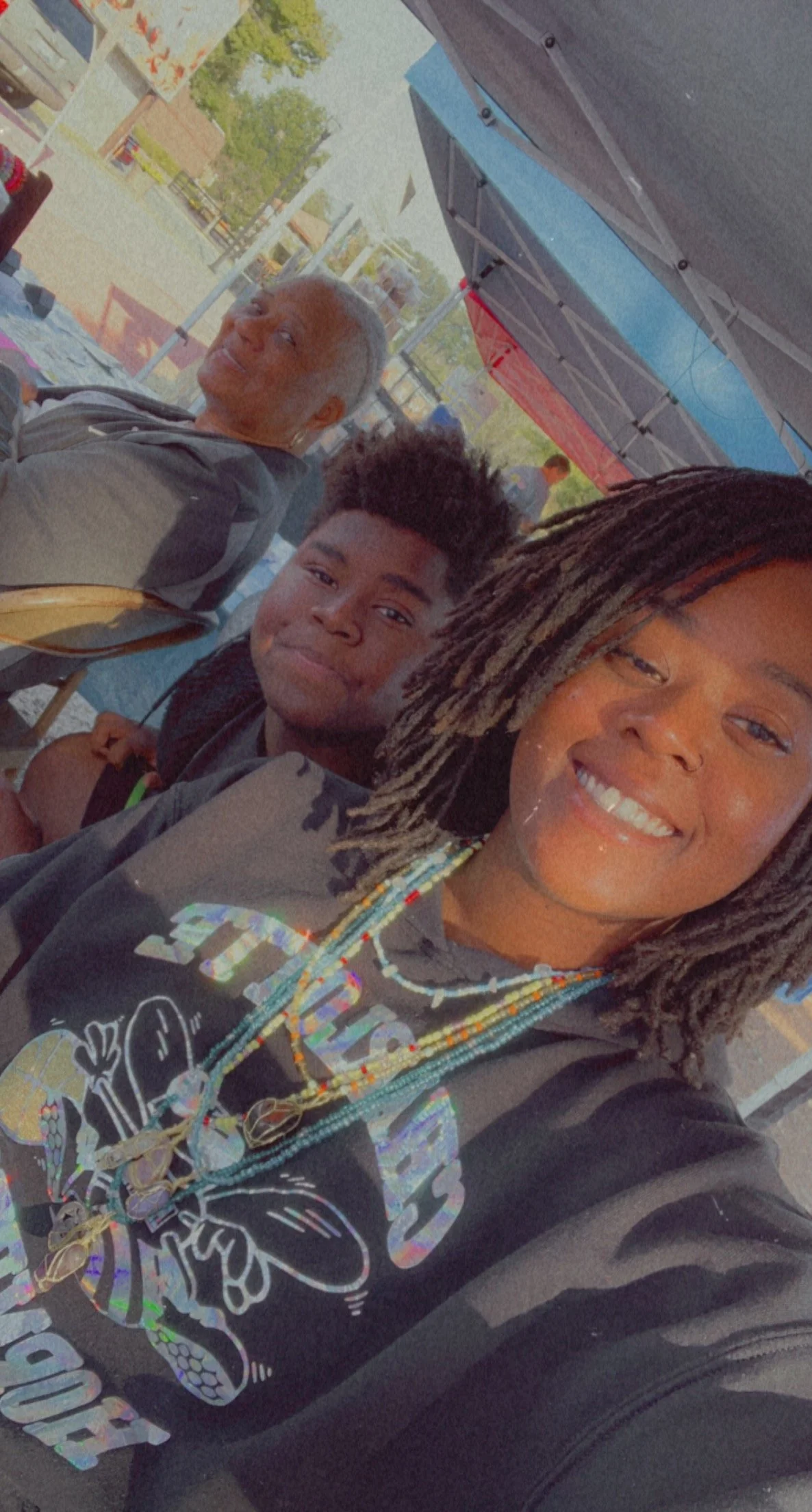 A woman with braided hair taking a selfie with two young people outdoors, standing near tents with a clear sky and trees in the background.