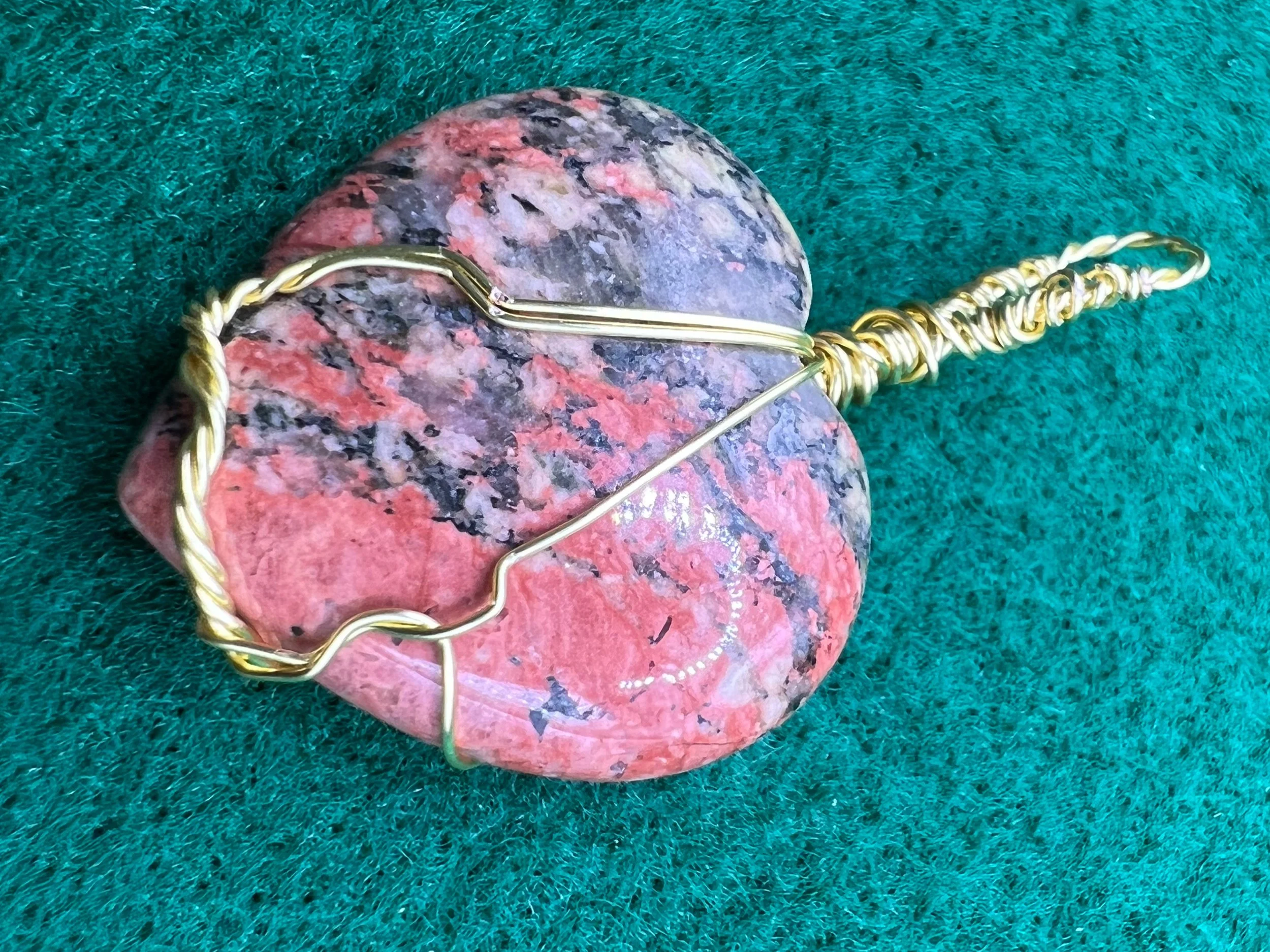 A colorful stone with pink, gray, and black hues lying on a green textured surface, with a gold-colored wire wrapped around it.