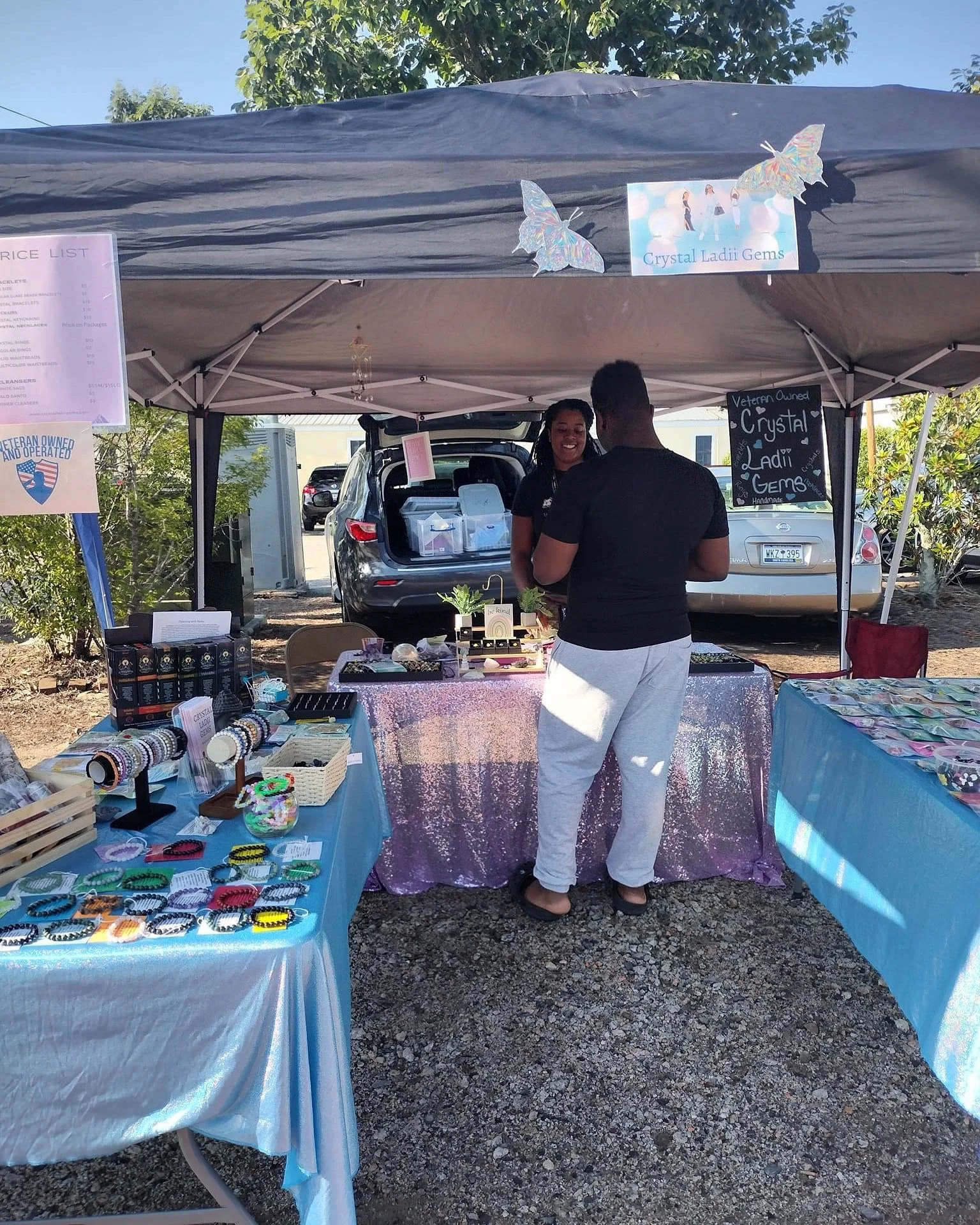 Outdoor market stall with jewelry and accessories on display tables, two women interacting, and a sign indicating handmade jewelry called 'Crystal Ladii Gems' with decorative butterfly and cloud decorations hanging from the tent.