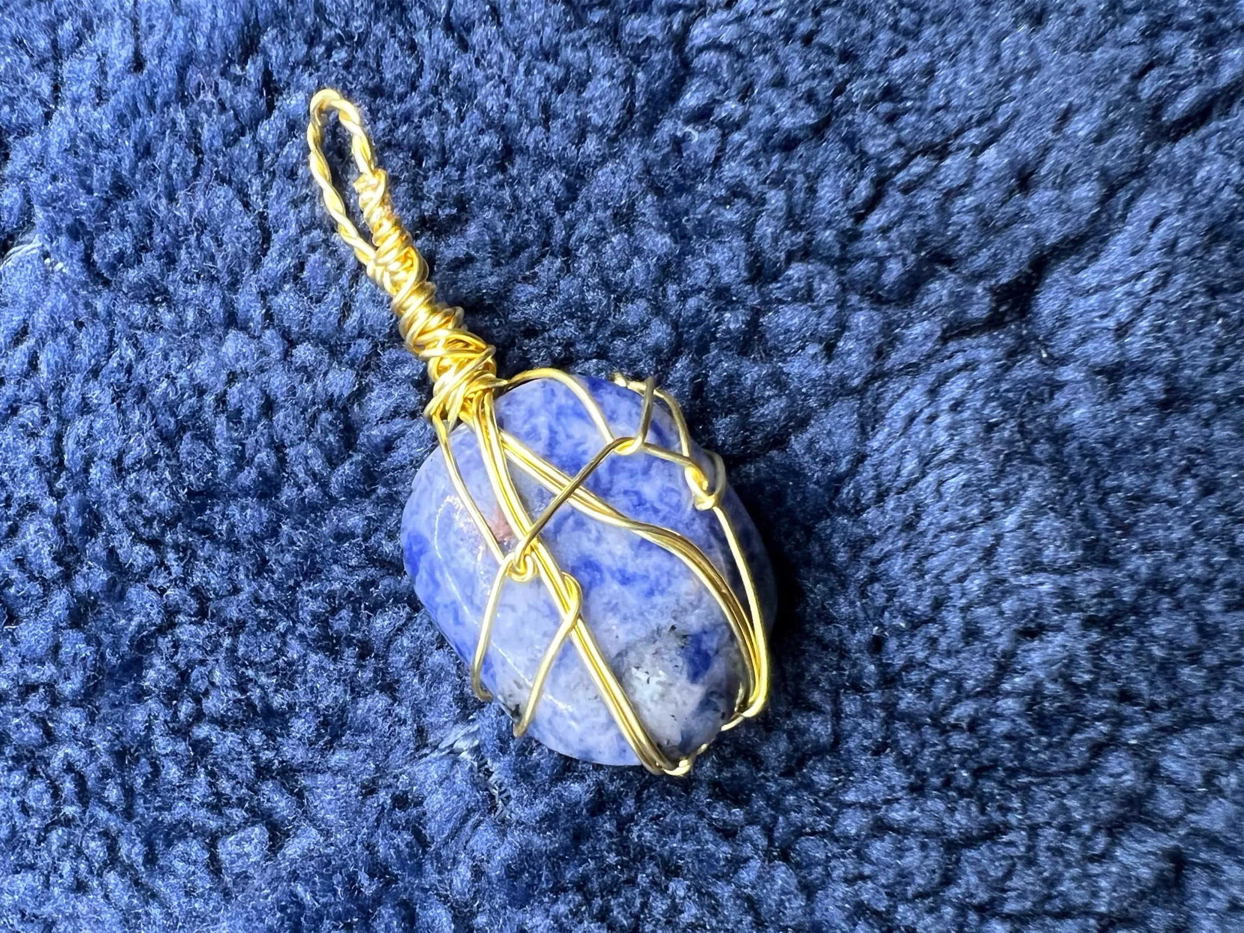 A teardrop-shaped blue stone wrapped in gold wire, resting on a dark textured fabric.