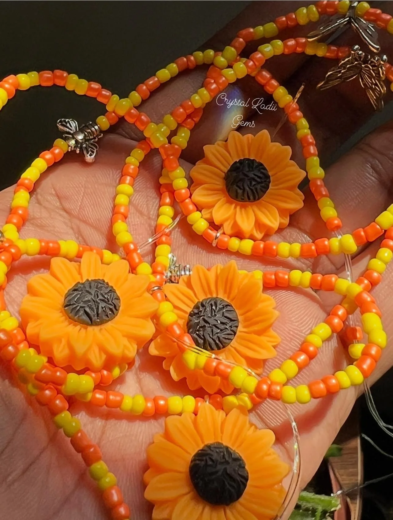 Close-up of orange sunflower-shaped earrings with black centers, yellow and orange beaded necklaces, and metal bee and butterfly charms, all placed on a person's hand.
