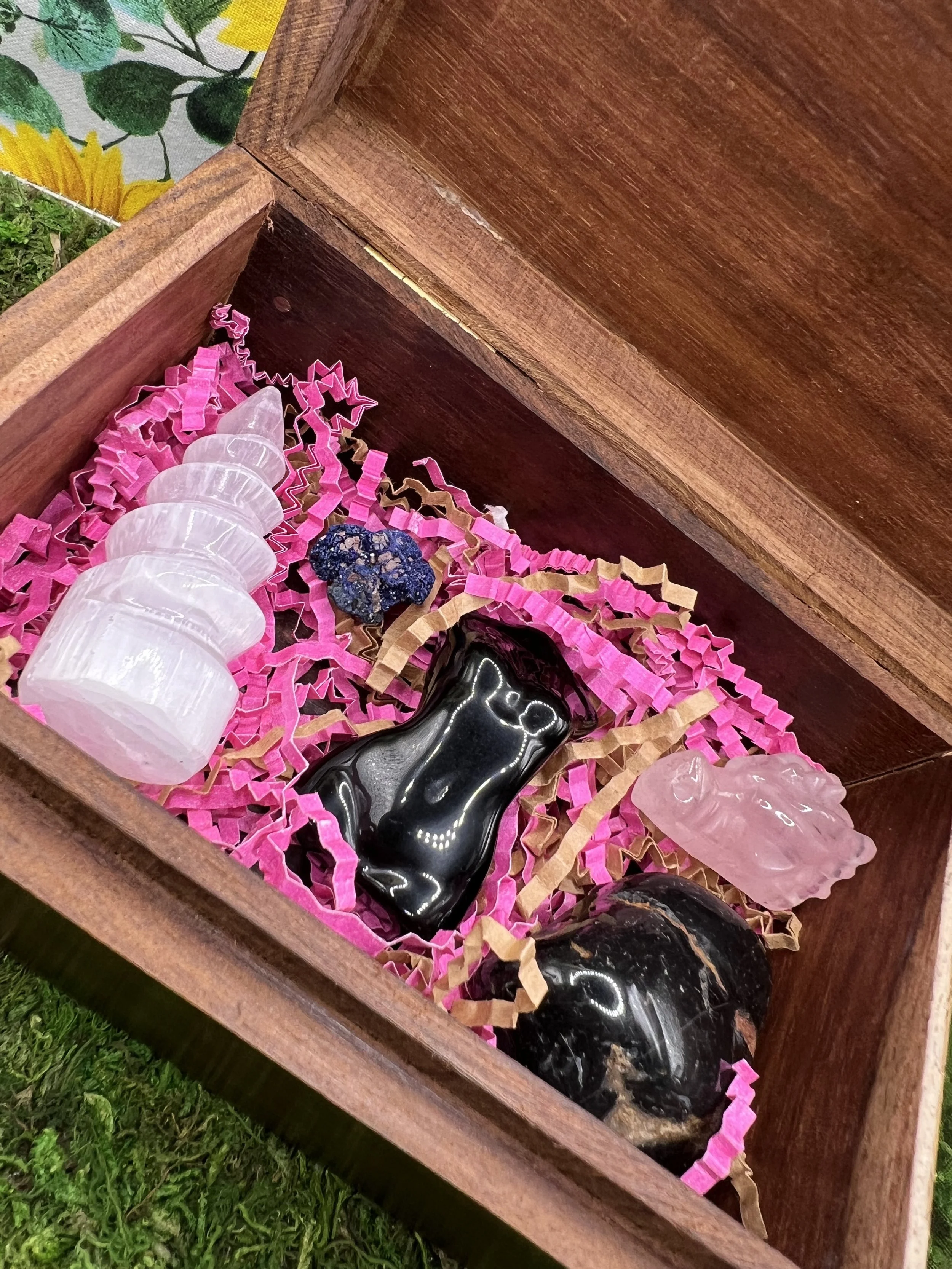 Close-up of a wooden box filled with pink paper shredded filler and black and pink animal-shaped items, with a green grassy area visible outside the box.