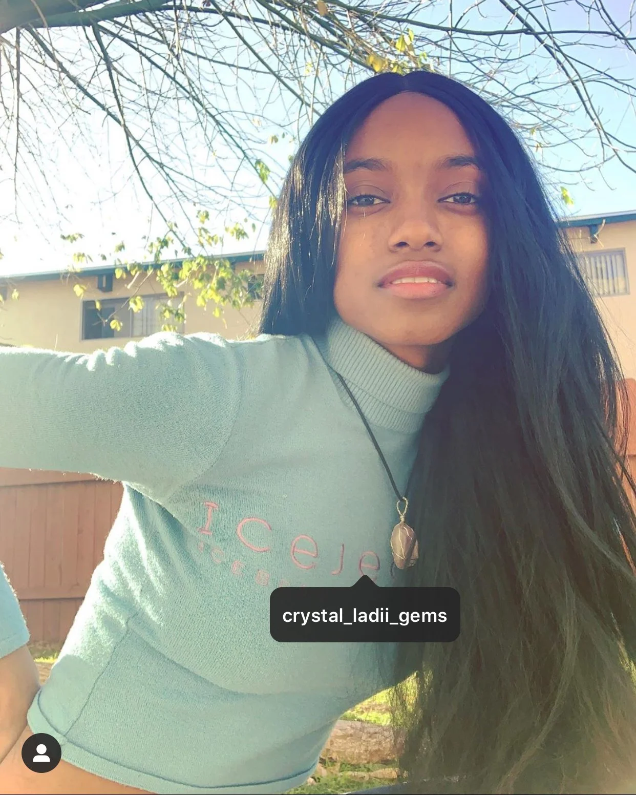 A young woman with long black hair and medium skin tone smiling outdoors on a sunny day. She wears a light blue sweater with pink lettering, a necklace with a large gemstone pendant, and is standing in front of a house with a wooden fence and leafles