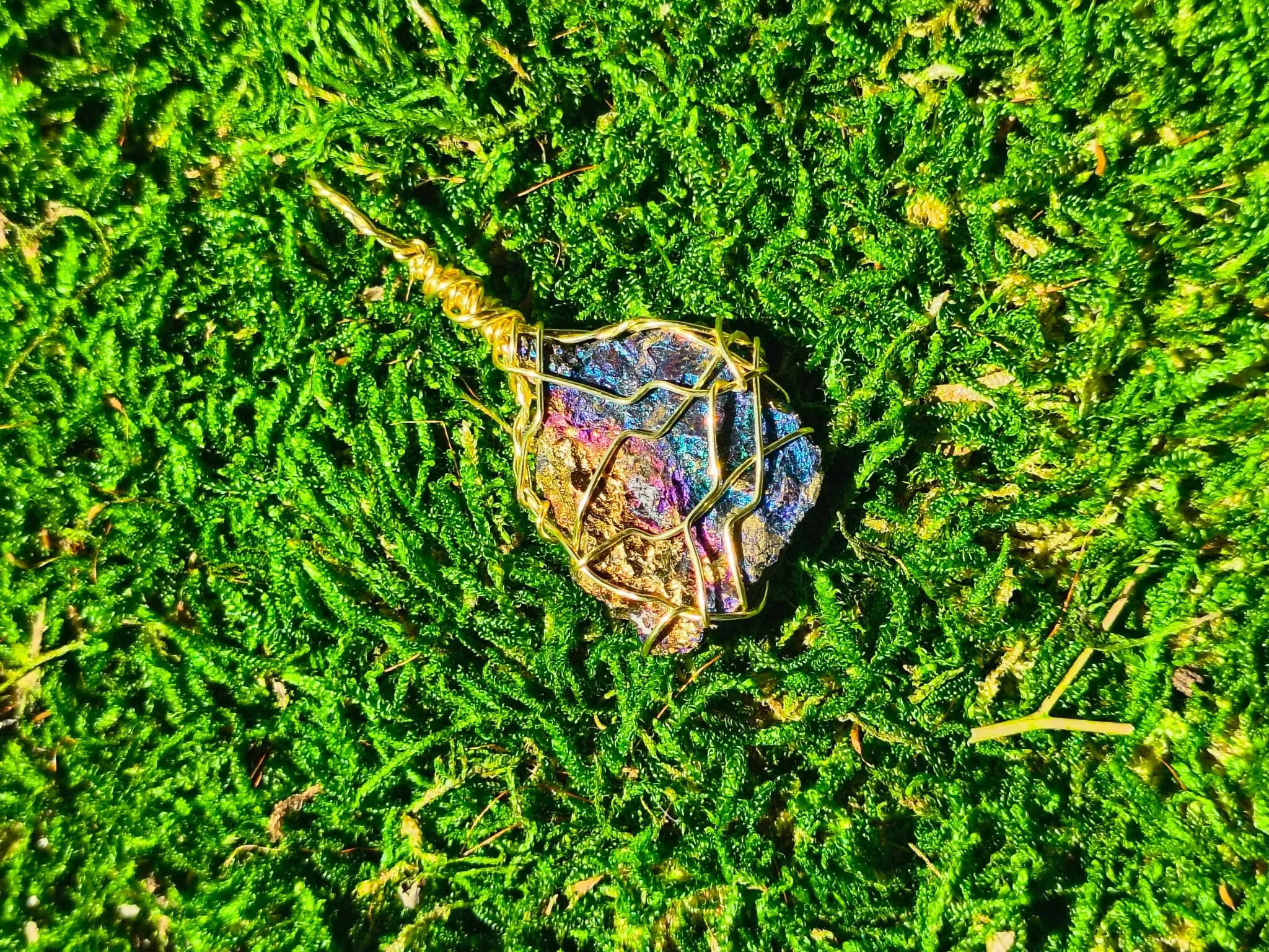 A colorful, iridescent gemstone wrapped in gold wire lying on green moss.