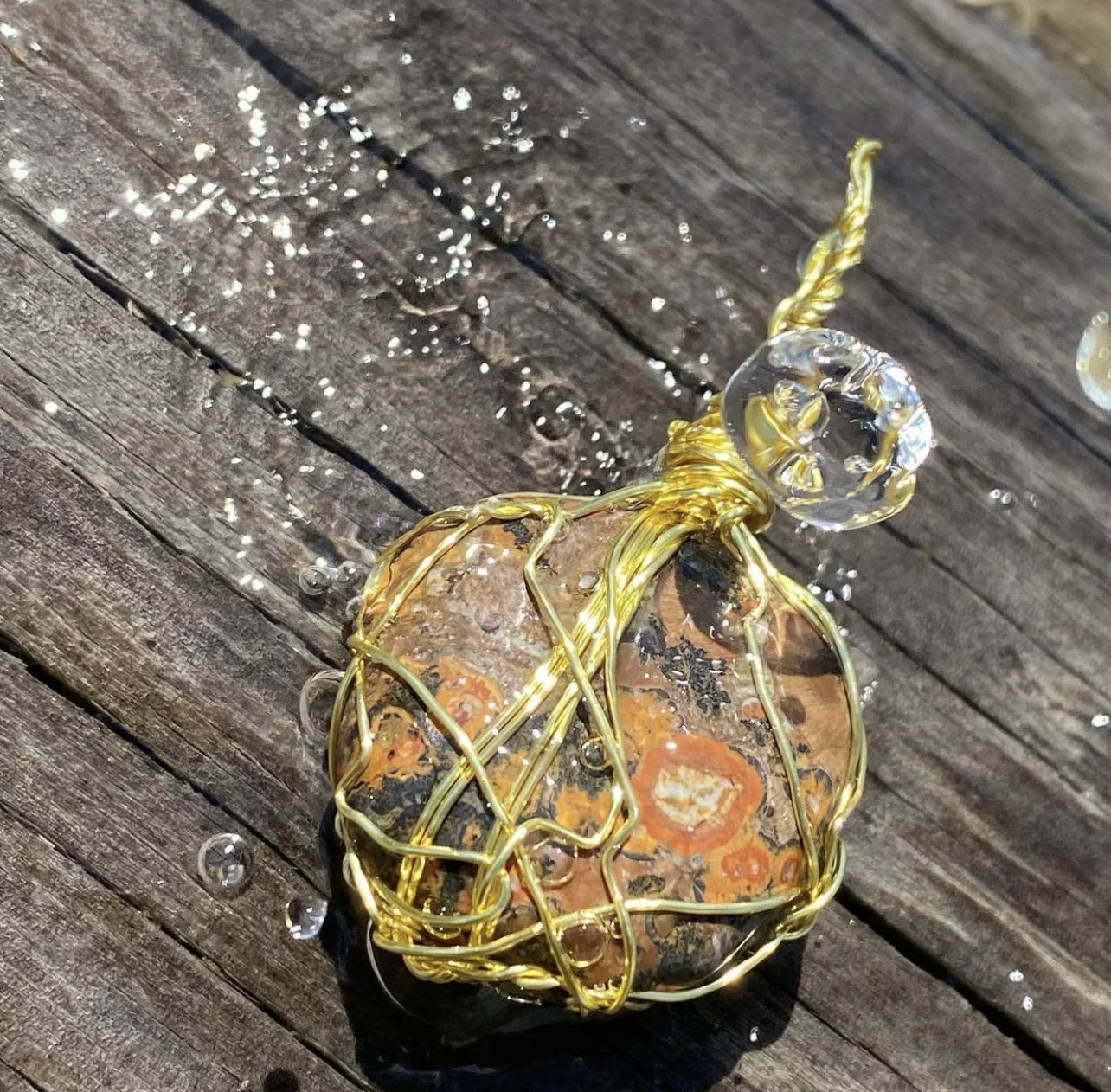 A decorative stone wrapped in gold wire with a clear circular glass ornament on top, placed on a wooden surface with water droplets around it.