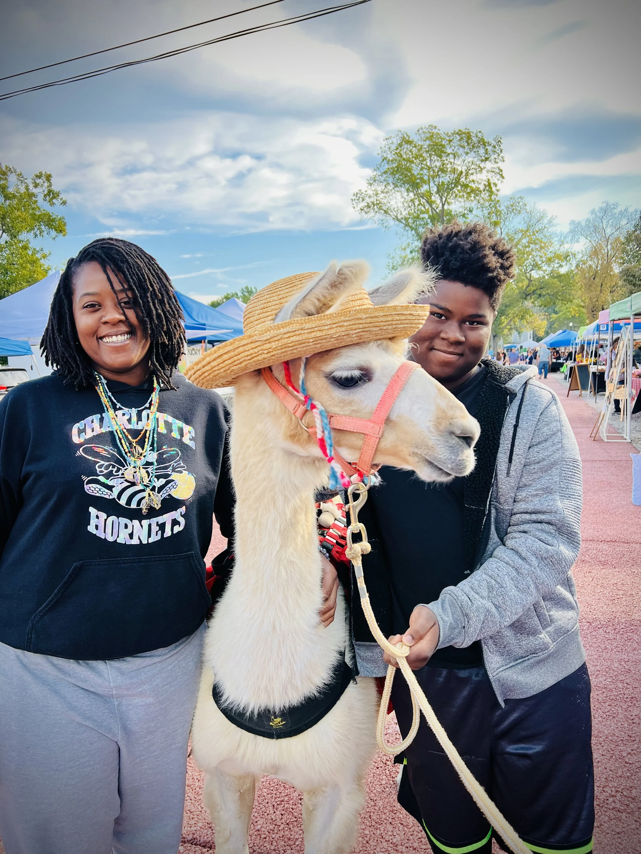 Two young people standing on either side of a llama decorated with a straw hat at an outdoor market with tents and trees in the background.