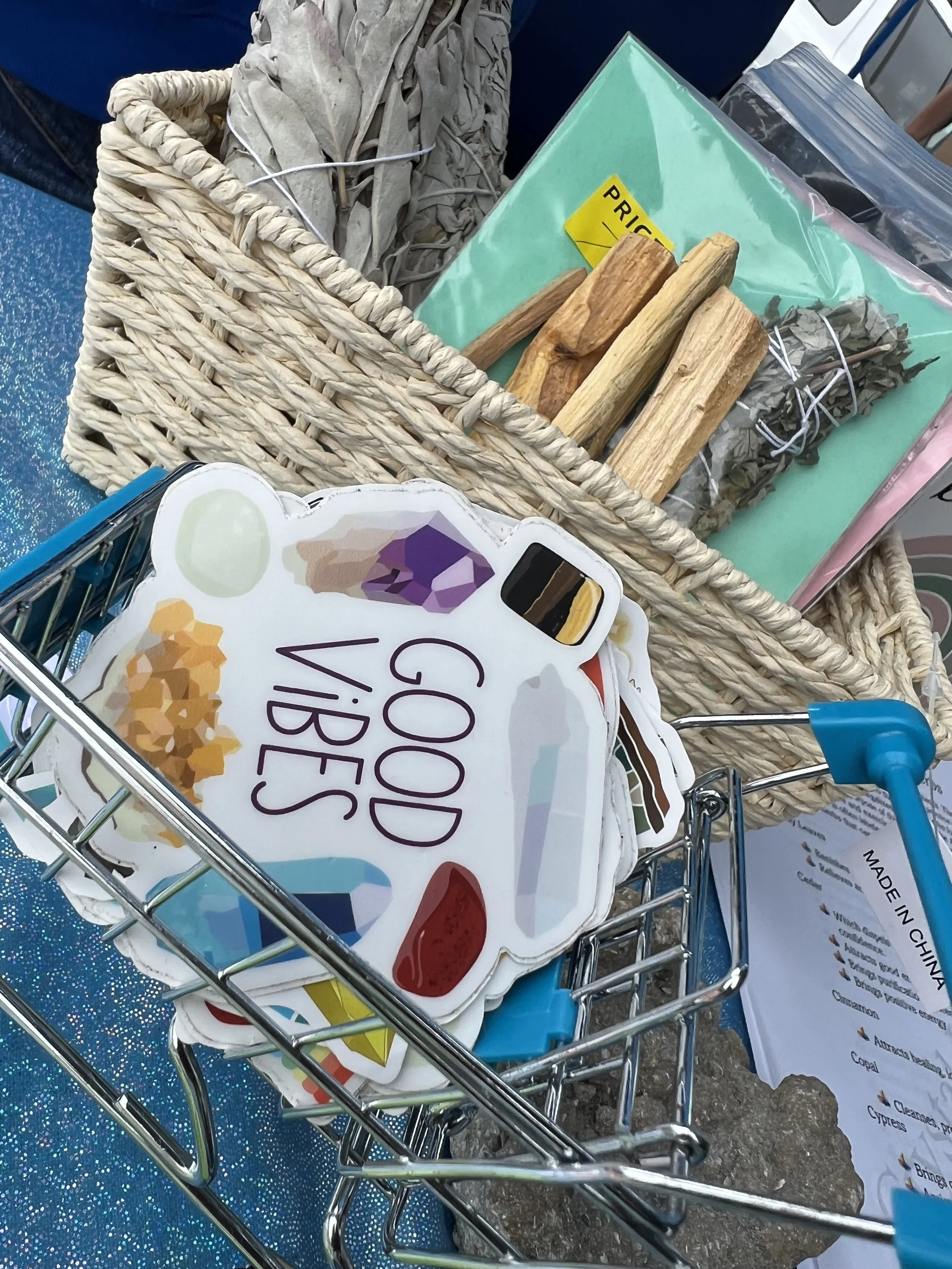 A shopping cart with a sign that says "Good Vibes" attached to it, containing a box of assorted cheeses, wooden sticks, and other grocery items at a store.