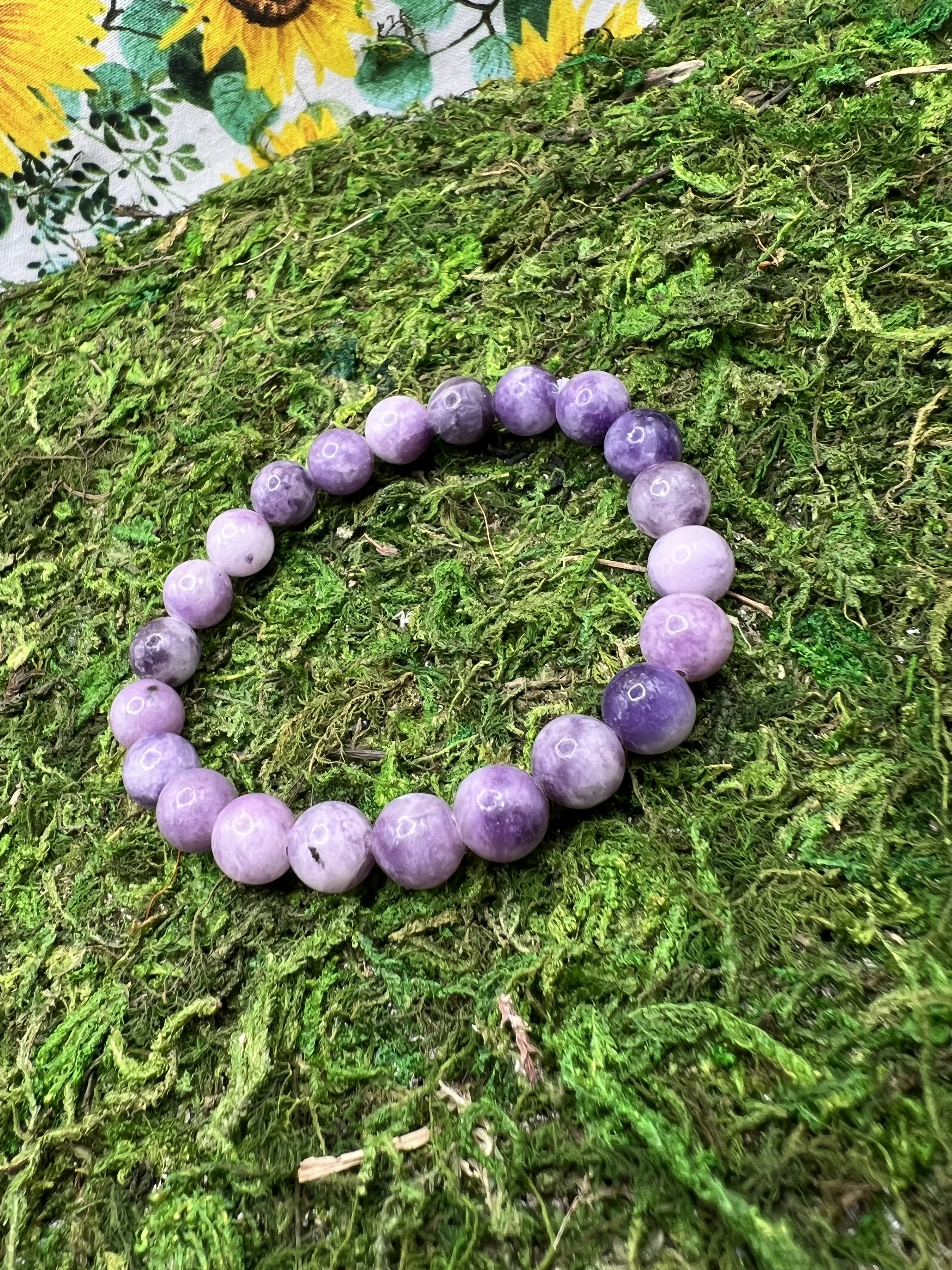 Purple beads arranged in a circle on a patch of green moss and twigs, with part of a sunflower-patterned fabric in the background.