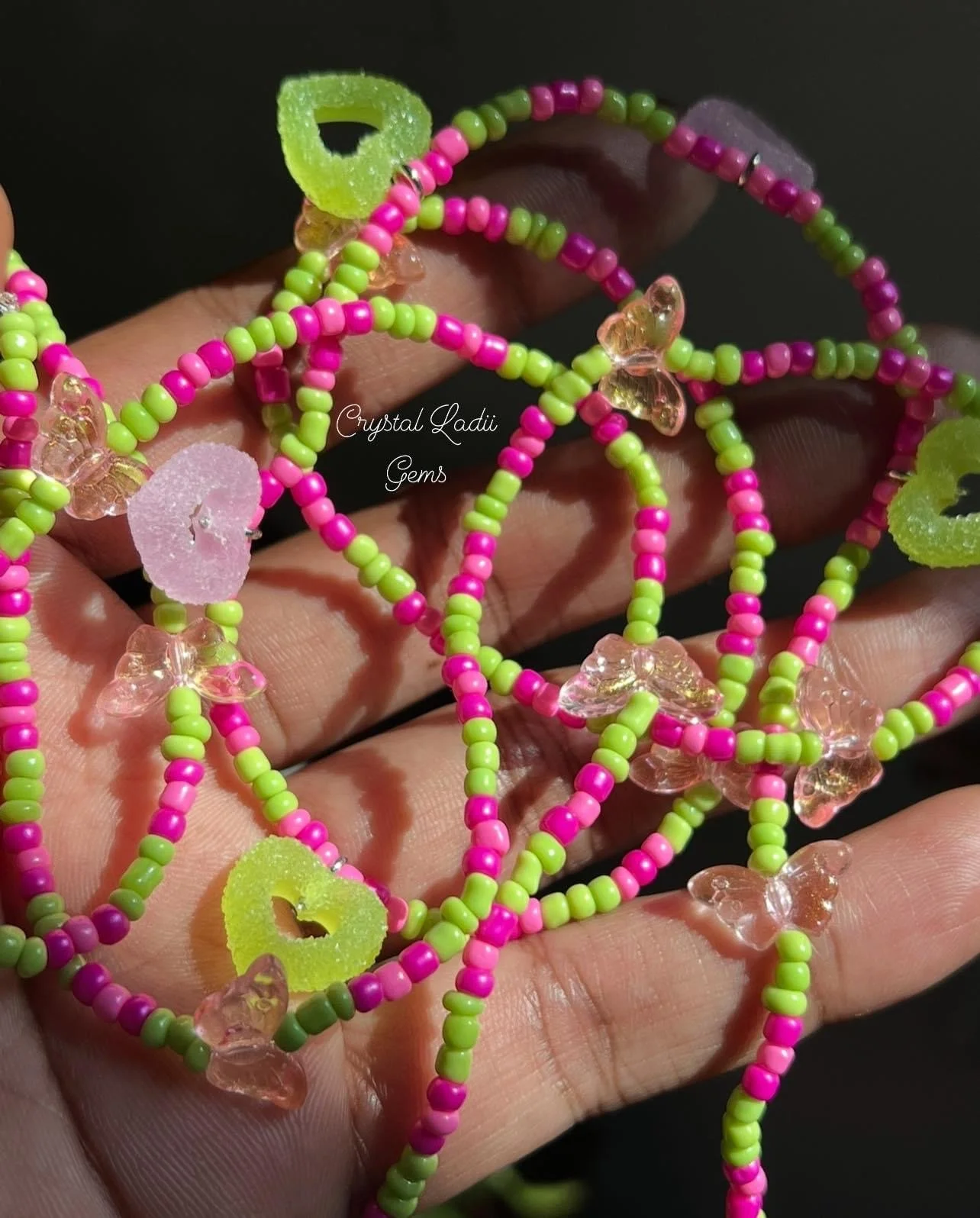 Close-up of colorful bead necklaces with pink, green, and transparent beads, some shaped like hearts and butterflies, being held in a person's hand.