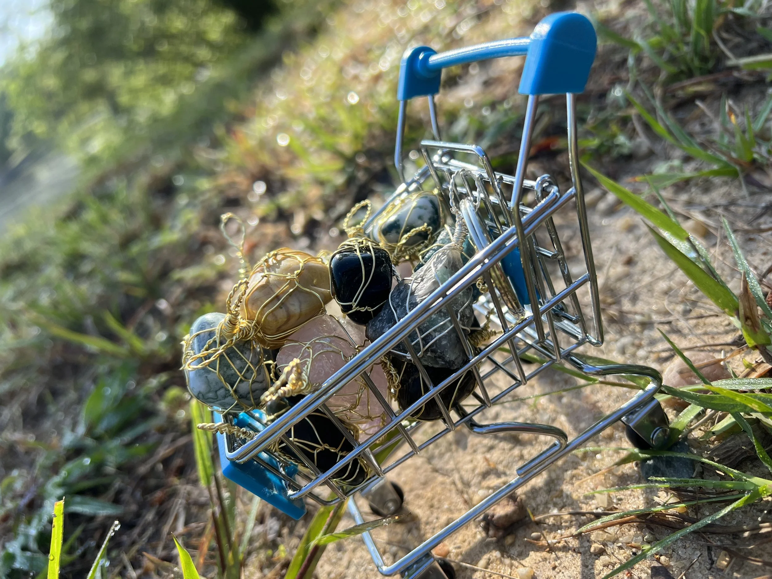 A small toy shopping cart filled with jewelry, including necklaces and bracelets, placed outdoors on the ground with grass and dirt.