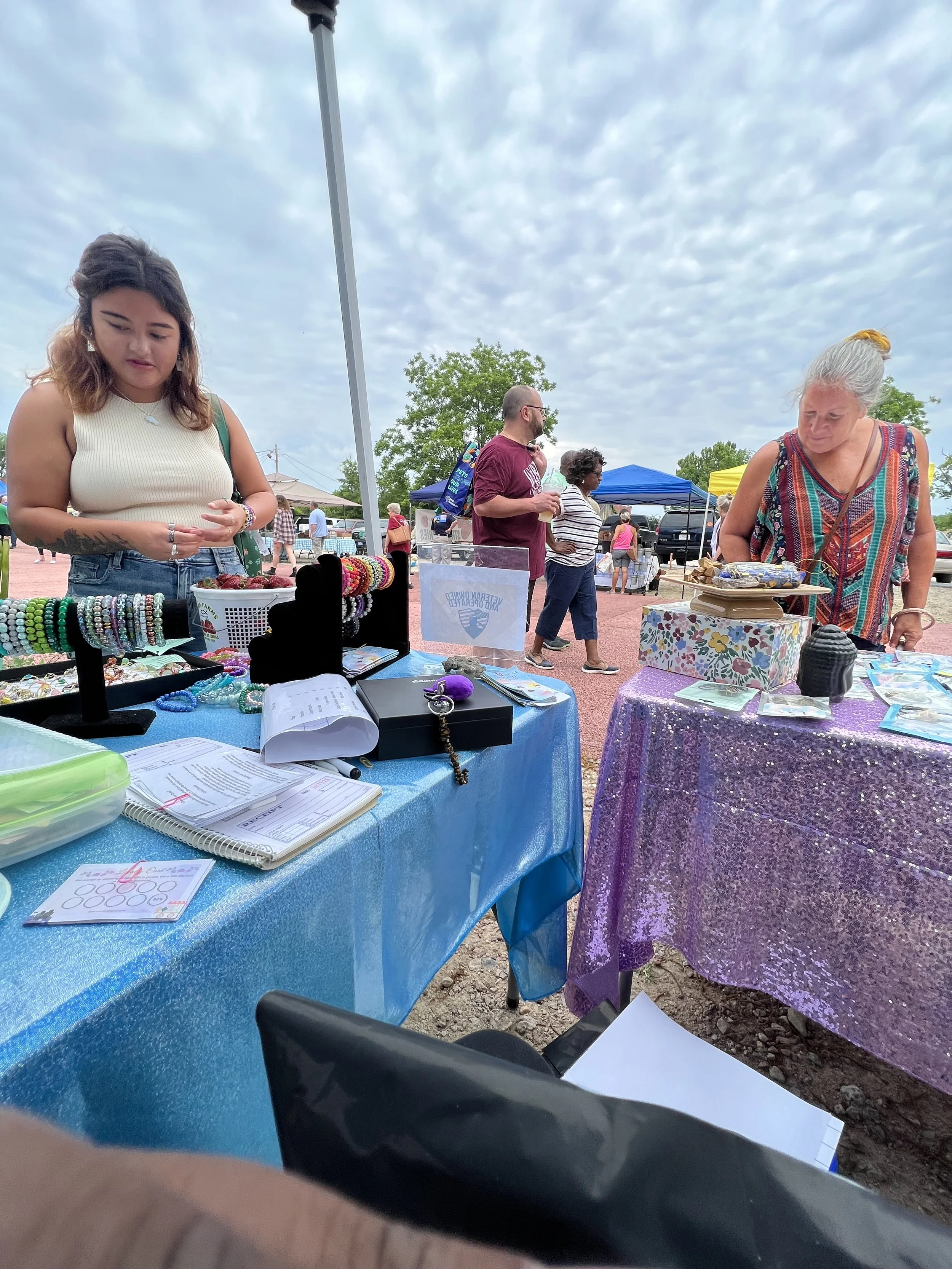 People browsing jewelry and small items at an outdoor market stall with tents set up in the background under partly cloudy skies.