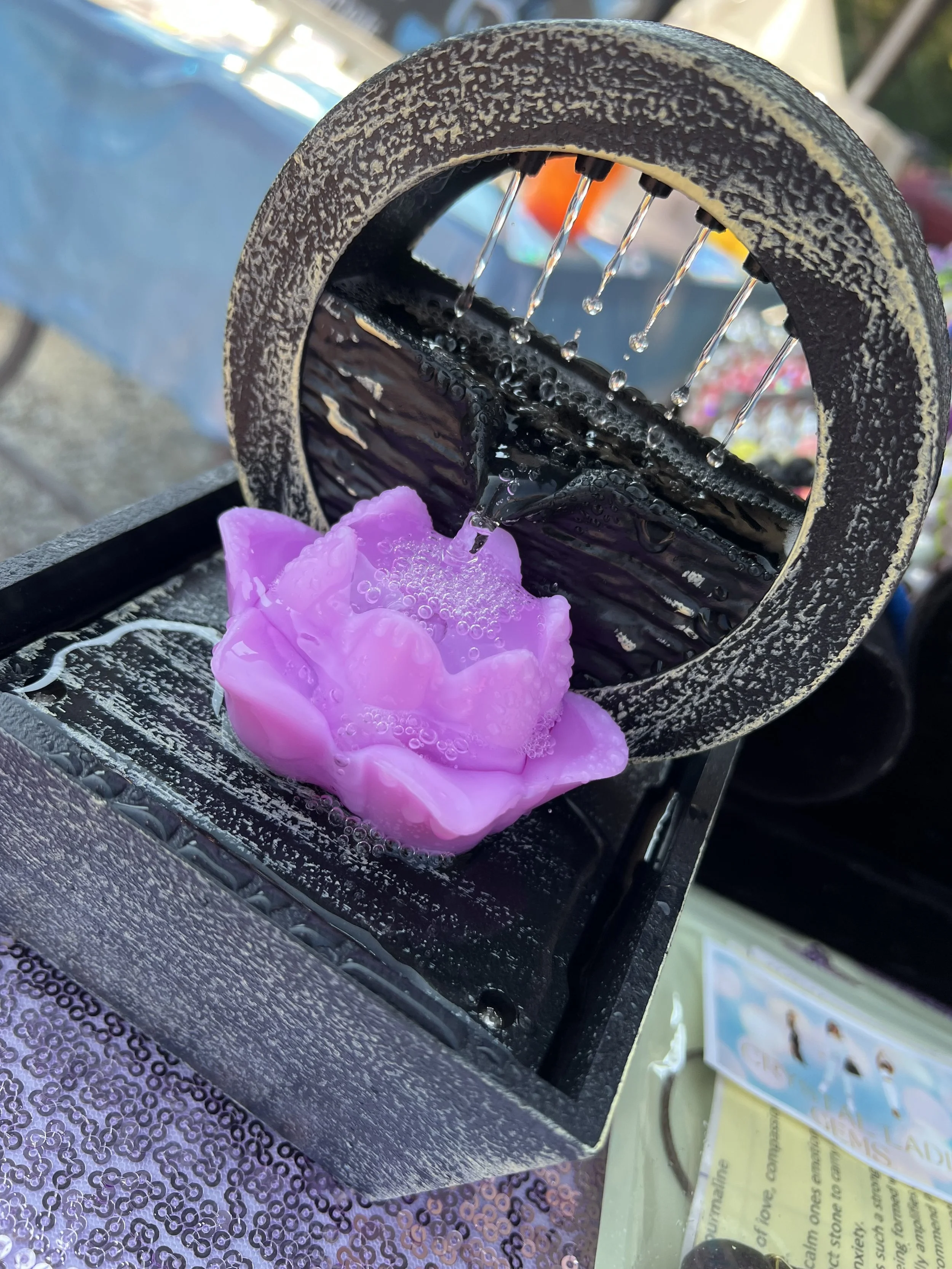 Close-up of a black handheld water pump with a pink soap or foam suds in front of a sequin-covered purple surface. Background shows what appears to be posters and outdoor scenery.