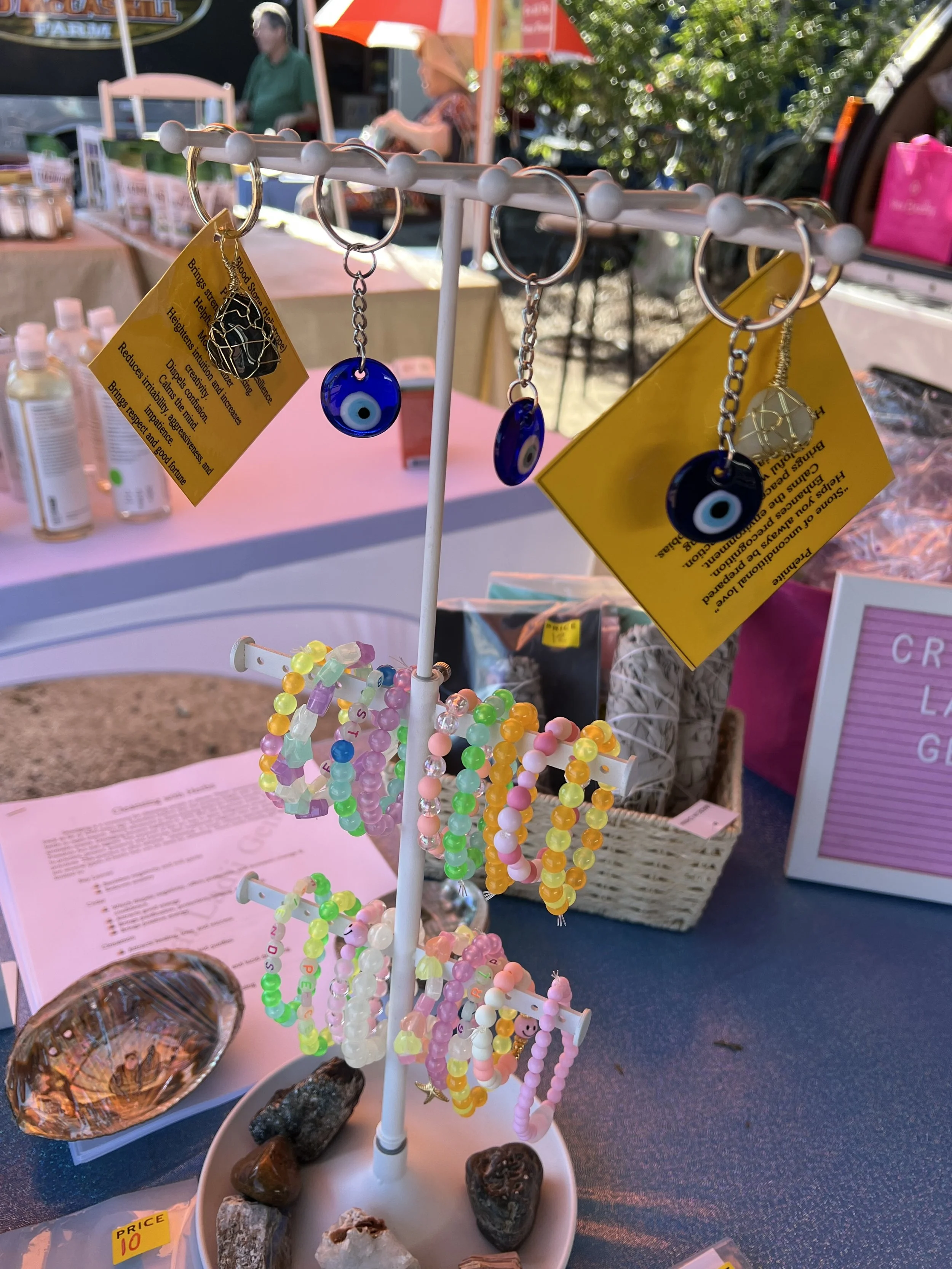 Colorful beaded bracelets, some with alphabet beads, hanging on a white display stand at an outdoor market. Stones and rocks are placed at the base of the stand. In the background, blurred vendors and market stalls can be seen.