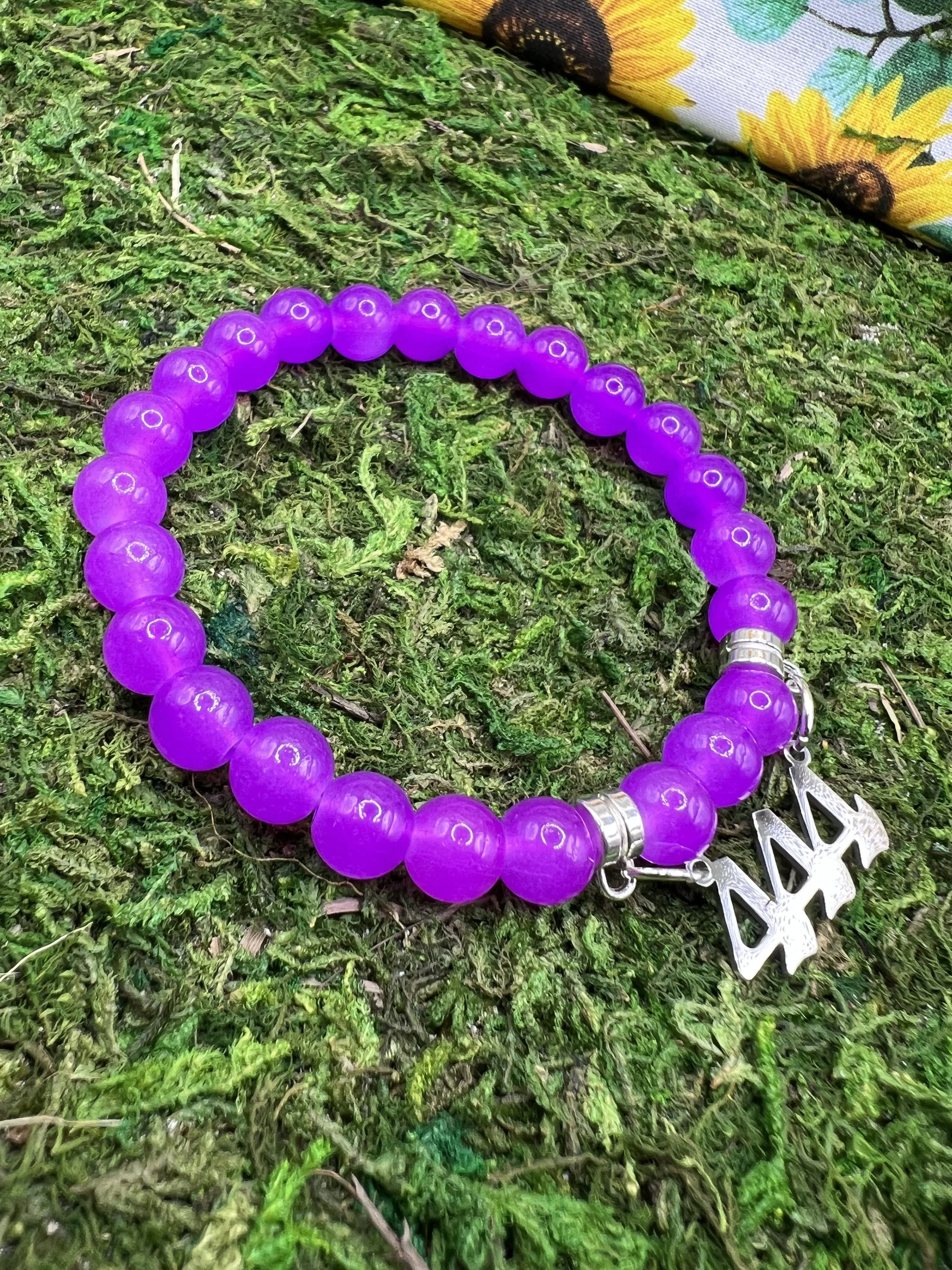 Close-up of a purple beaded bracelet with a silver snowflake charm lying on green moss with a piece of colorful fabric with sunflower and sunflower seed design in the background.