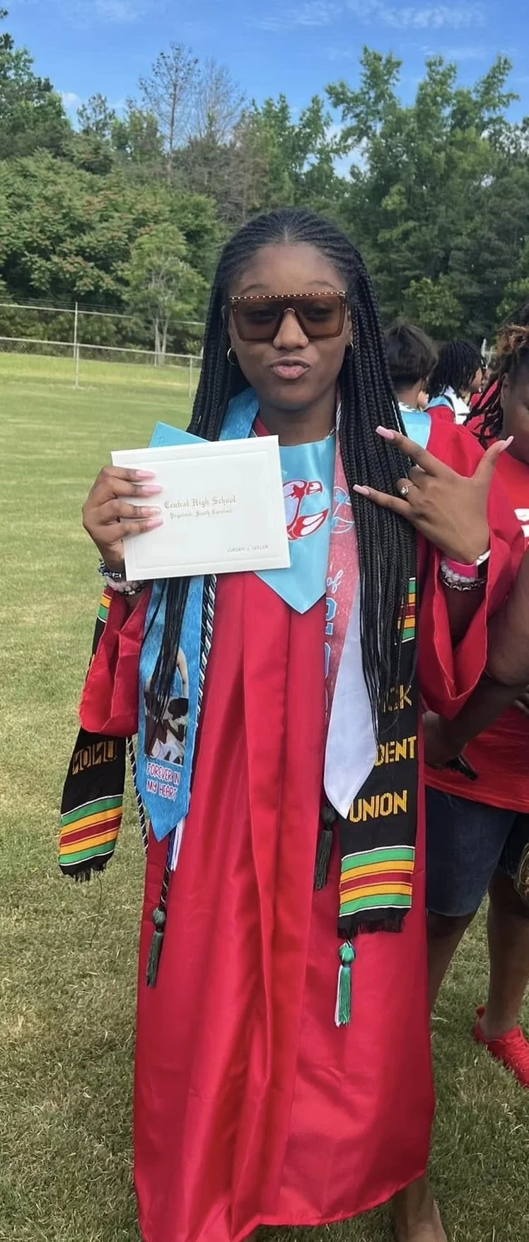 Young woman in graduation gown holding a diploma, wearing sunglasses and making a peace sign, outdoors on a grassy field.