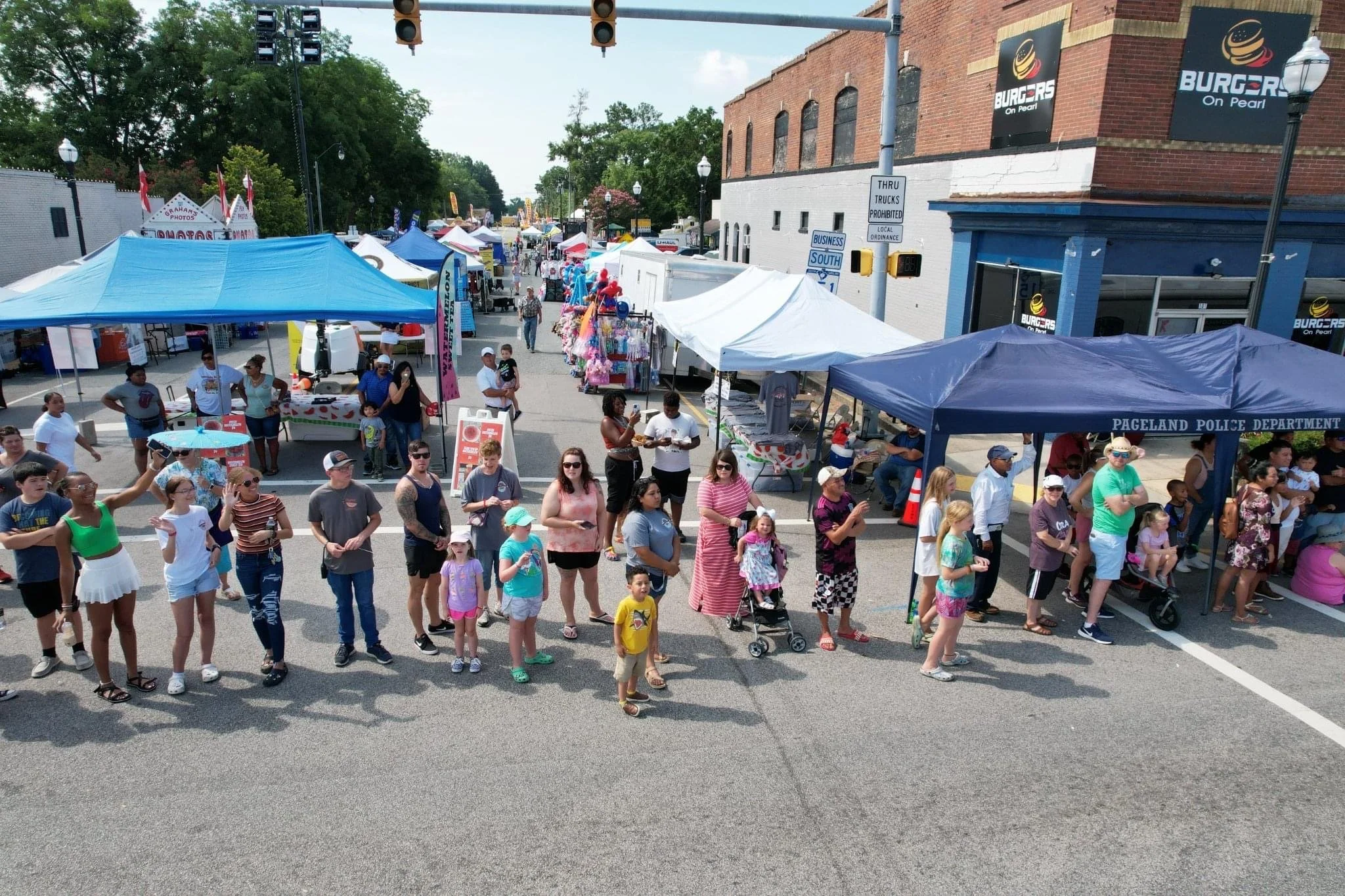 A street fair with a crowd of people, tents, and vendor booths. Some people are standing, walking, and chatting, while others are sitting or pushing strollers. Several tents and signs are visible, including one that reads "Pageland Police Department." The background shows a brick building with signs for "Burgers on Pearl" and street signs.