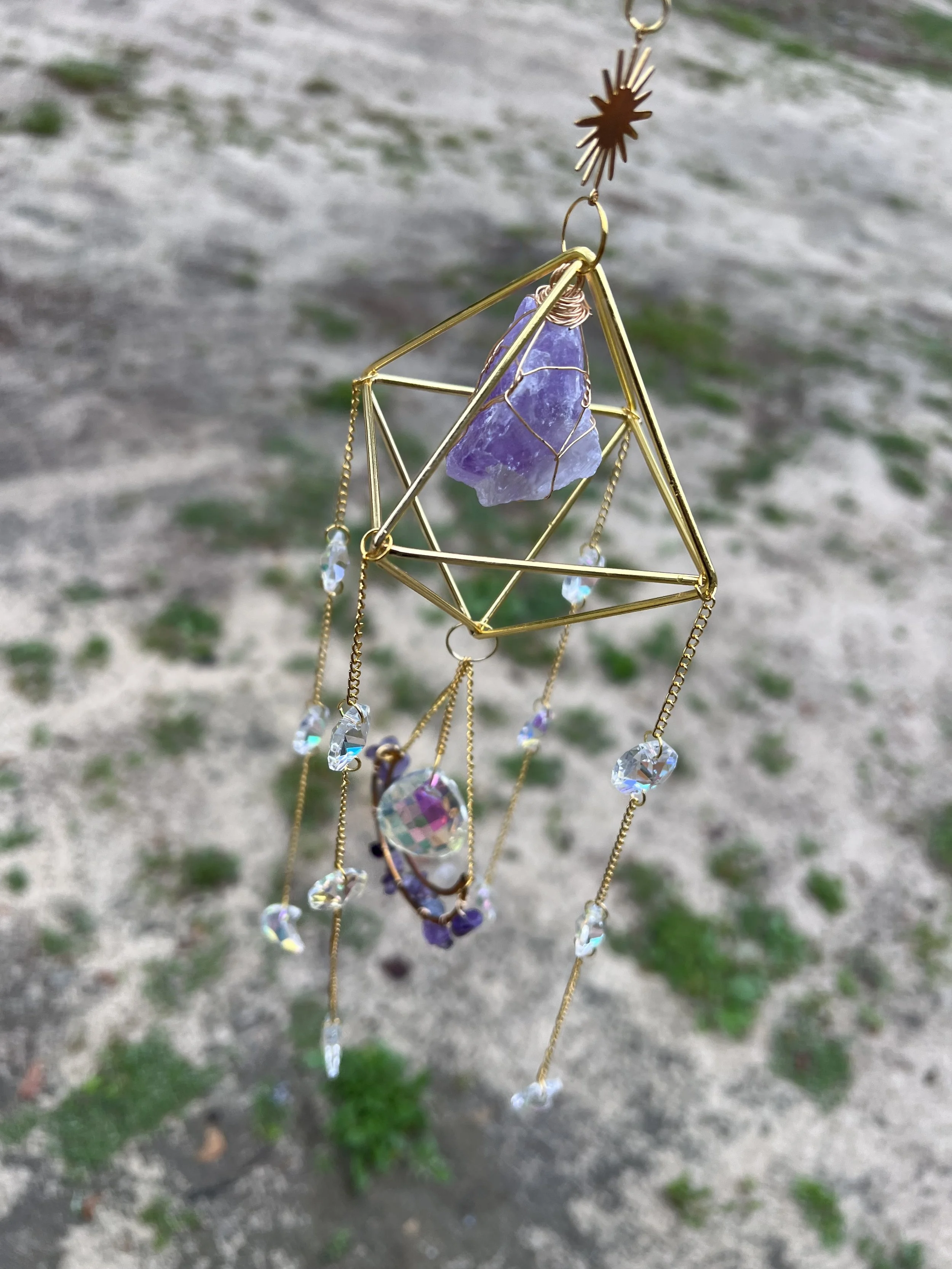 A gold geometric ornament with purple stone and hanging crystal beads, hanging outdoors over a dirt and grass background.