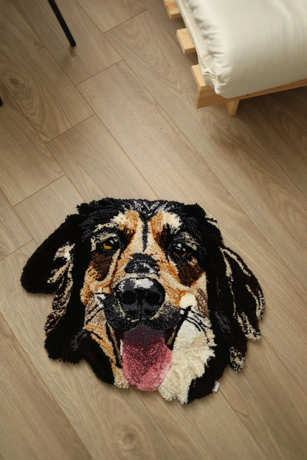 A rug in the shape of a dog's head with detailed facial features, fur markings, and a sticking out pink tongue, placed on a wooden floor near a bed.