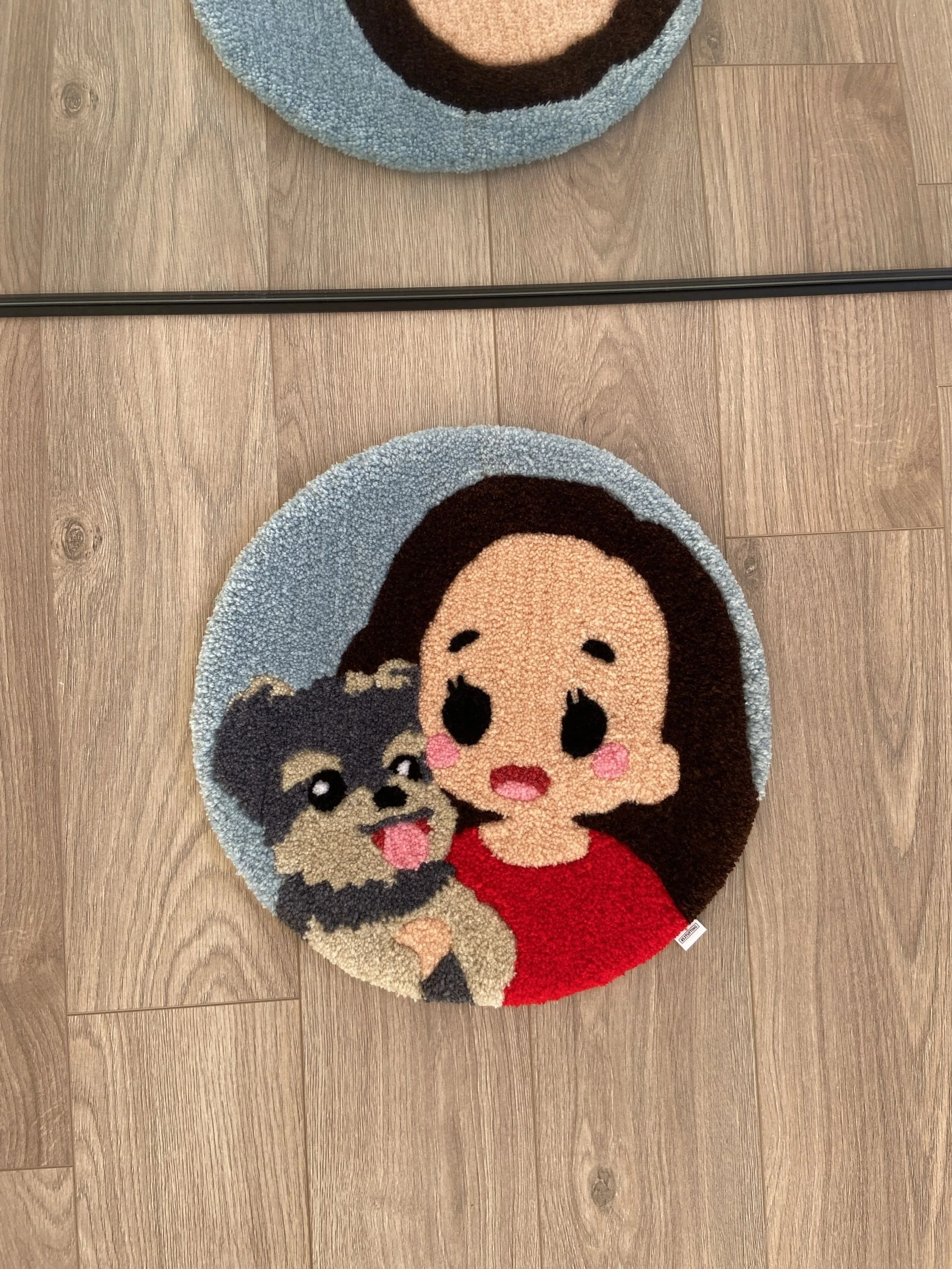 A round rug with a cartoon girl character, dark hair, rosy cheeks, and a red dress, on a wooden floor.