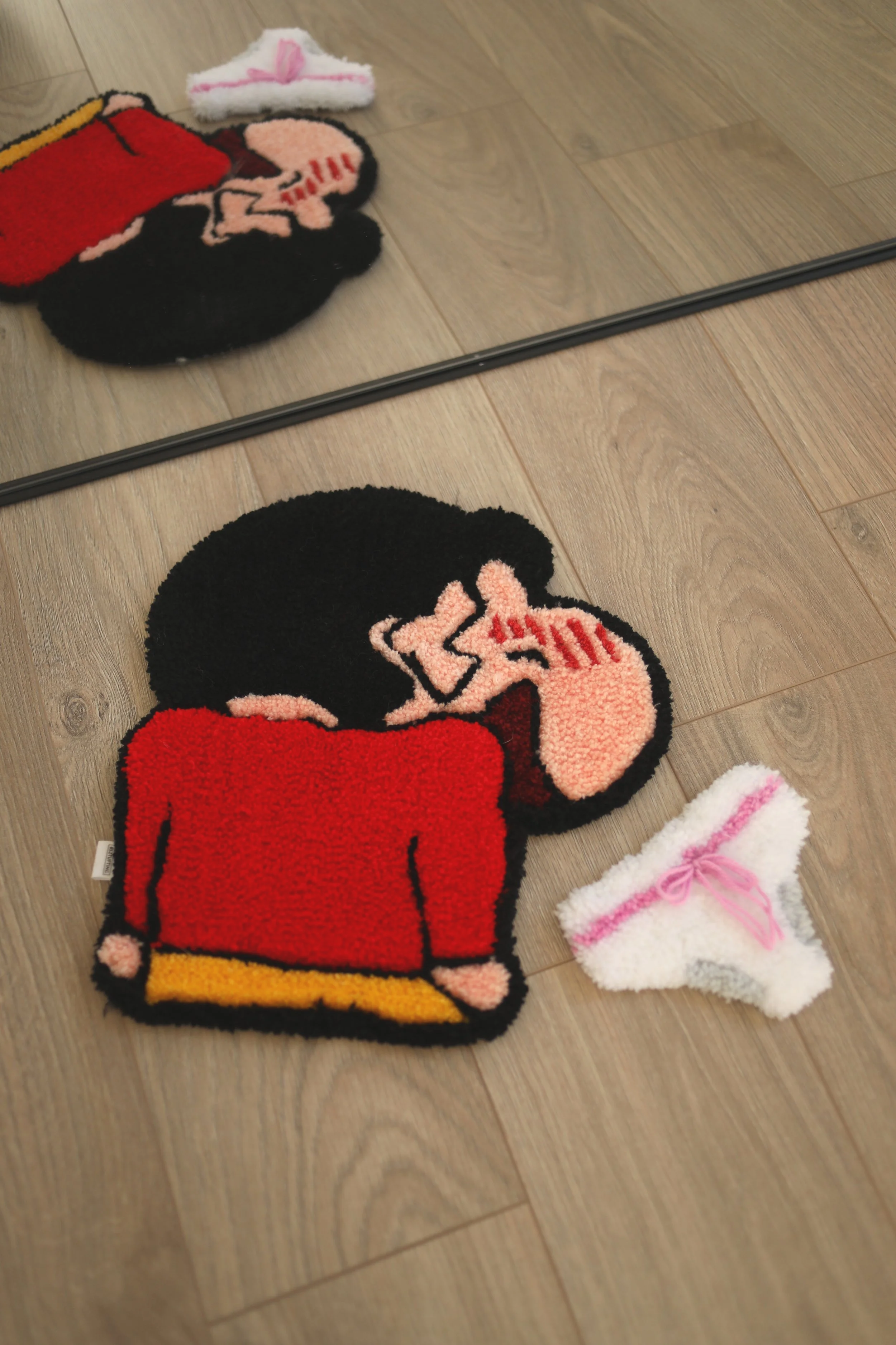 A pair of fuzzy slippers shaped like Mickey Mouse, with a red shirt, yellow shoes, and black head, placed on a wooden floor near a mirror. A small pink and white fluffy Christmas stocking with a pink ribbon is partially visible.