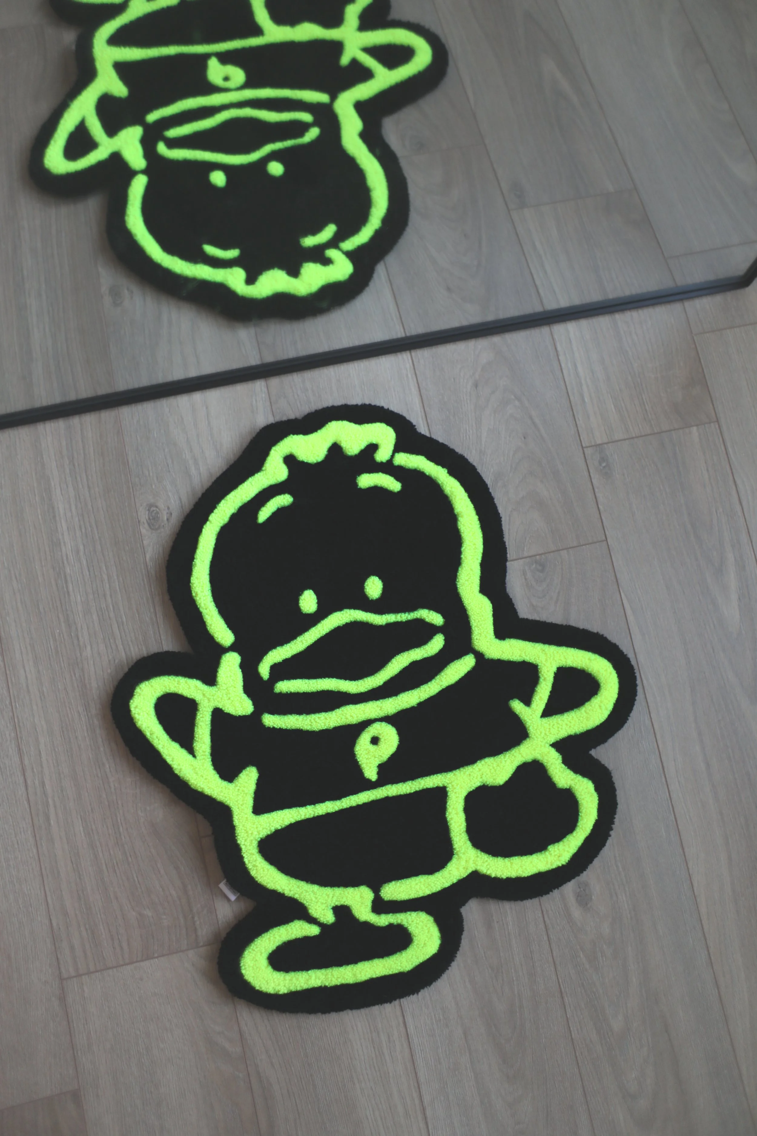 A black rug with neon green outline of comic-style duck character with a pink beak, yellow eyes, and a duck's bill in a speech bubble, placed on a wooden floor.