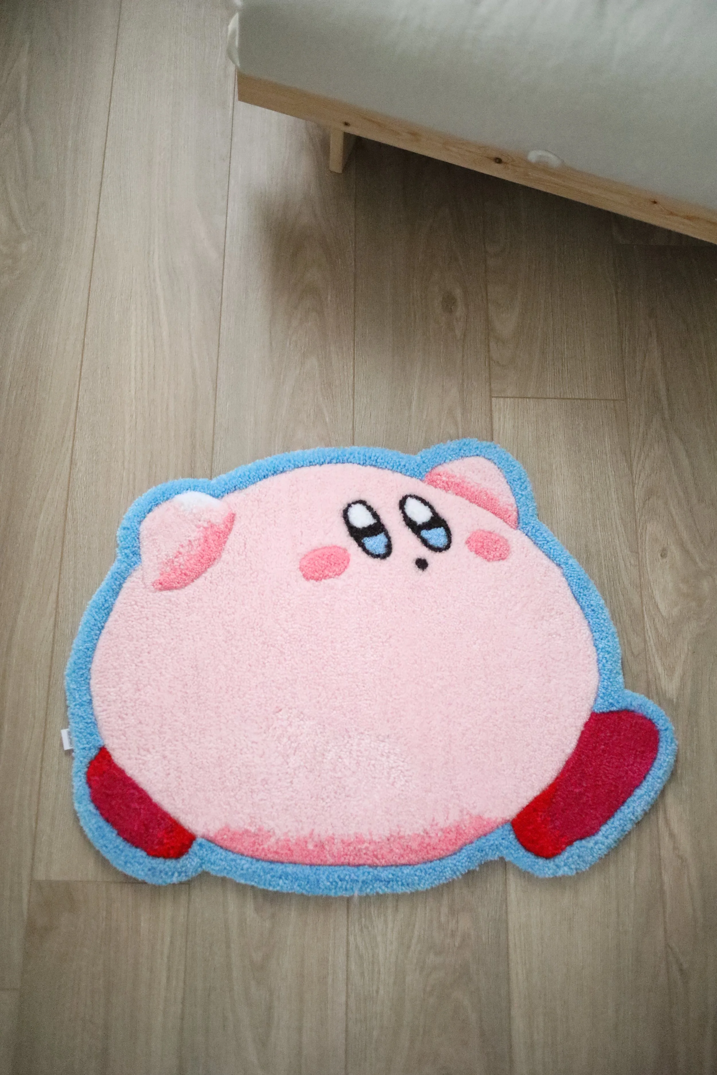 A plush rug designed to look like Kirby, a pink video game character, with blue eyes and a cute facial expression, placed on a wooden floor.