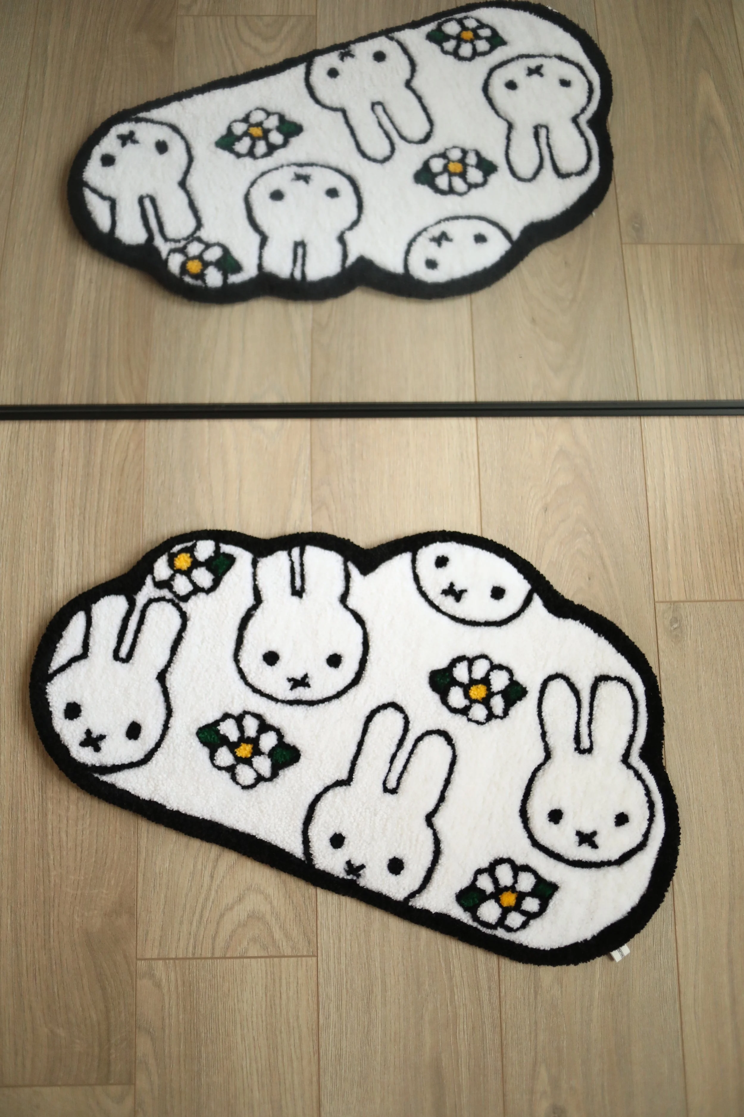 A white oval-shaped area rug with a black border, featuring cartoon bunny rabbits and white, black, yellow, and green floral patterns, placed on a wooden floor.