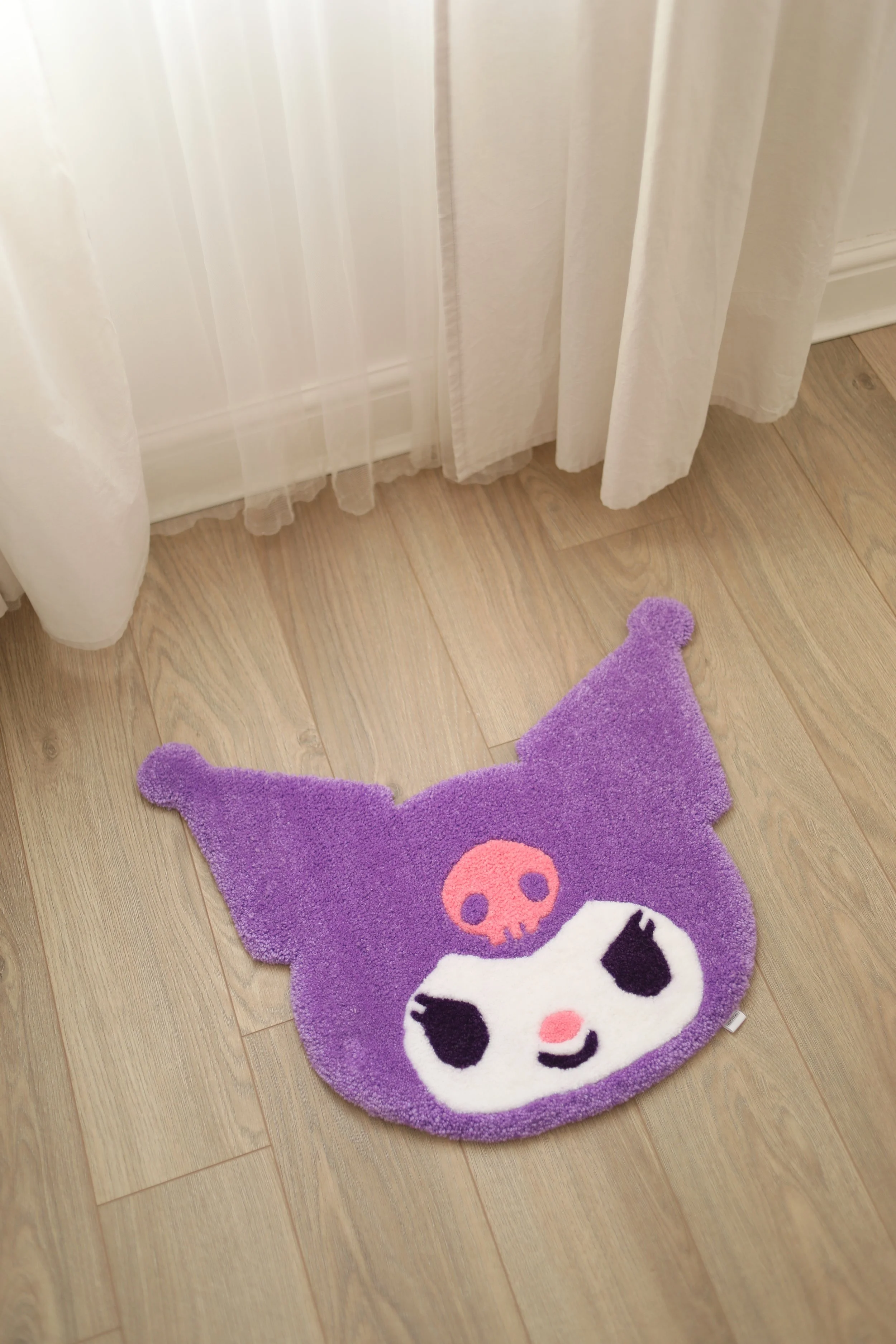 A purple plush rug shaped like a skull and crossbones lying on a wooden floor, with white curtains hanging nearby.