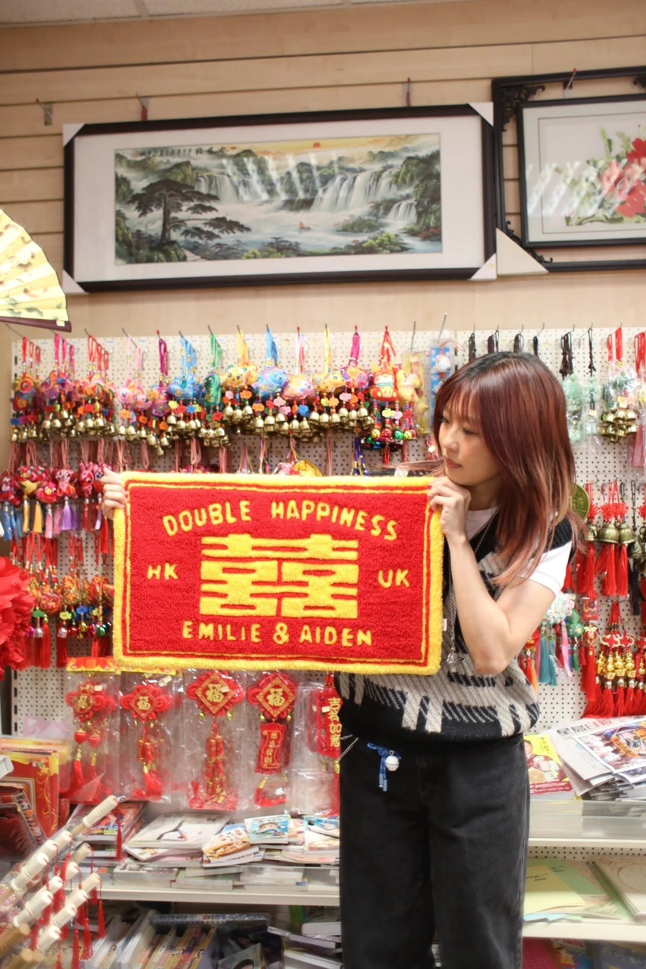 Woman in black pants and white and black top holding a red and yellow double happiness rug in a store decorated with Chinese New Year items.