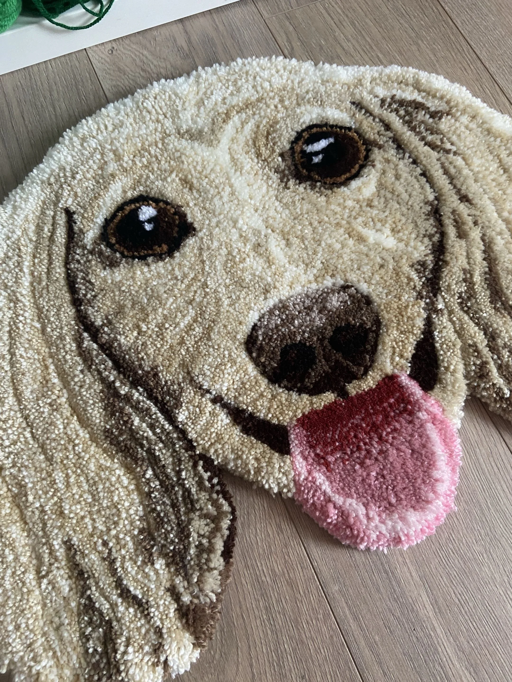 Close-up of a round pet rug with a cartoon dog face, featuring brown eyes, a brown nose, and a pink tongue, placed on a wooden floor.