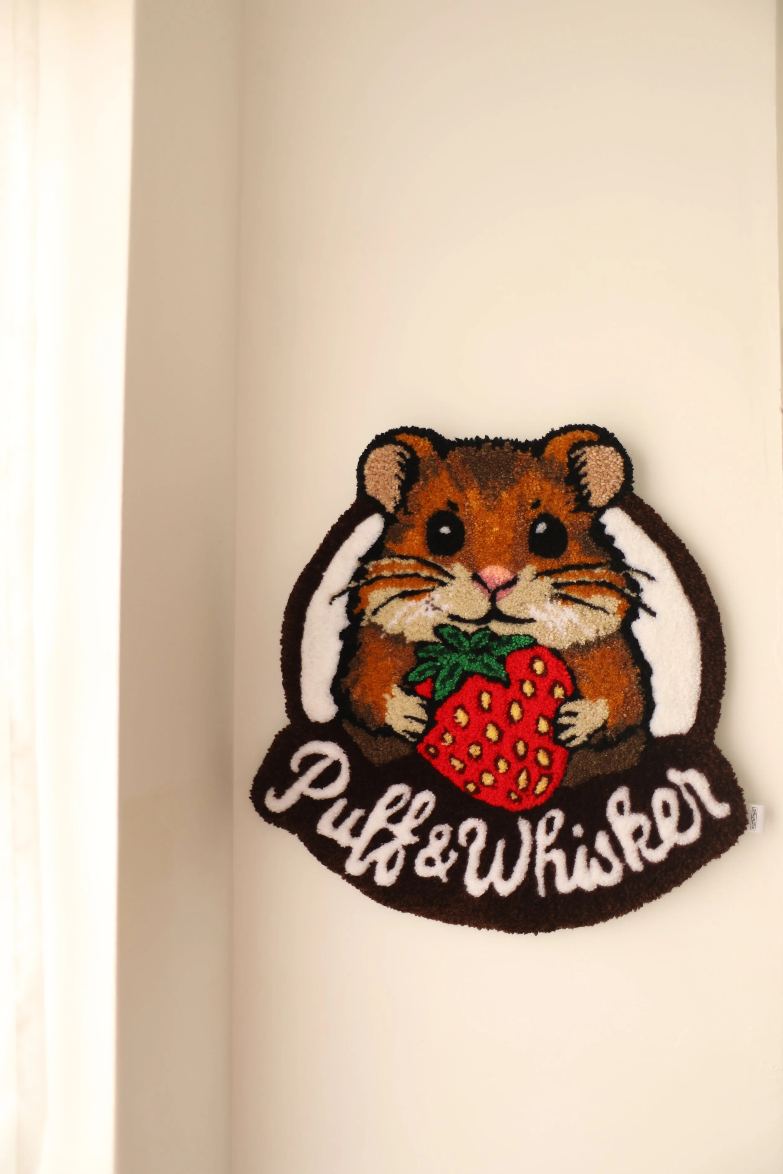 A decorative rug hanging on a white wall featuring a hamster holding a strawberry with green leaves. The rug has the text 'Puff & Wiser' at the bottom.