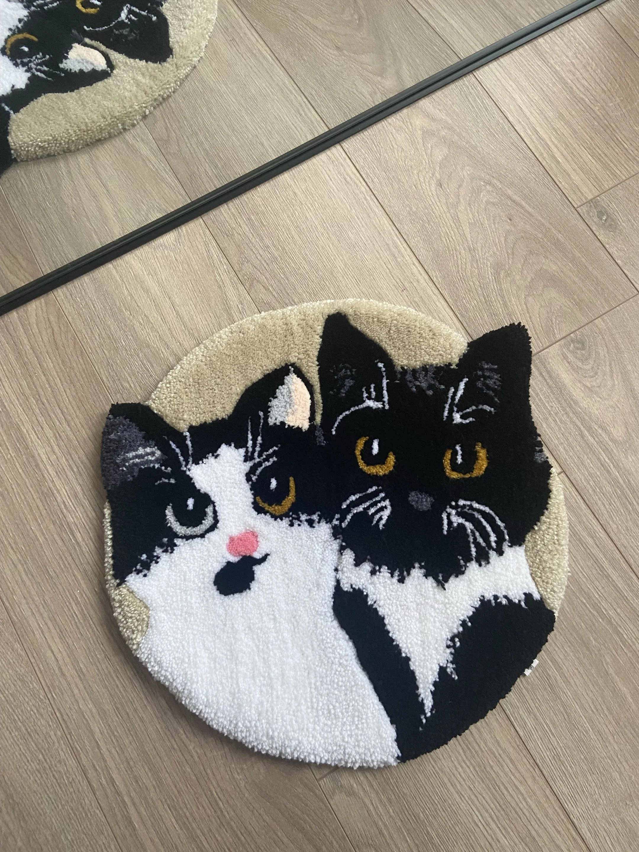 A circular rug with a black and white cat face design, pink nose, and yellow eyes, on a wooden floor near a mirror and black border.