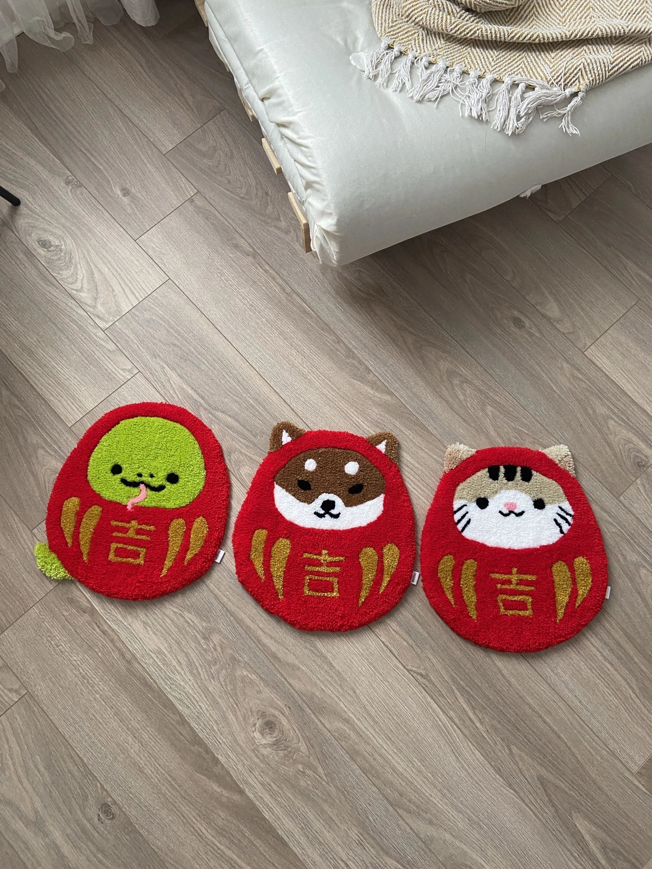 Three red daruma rug with animal and character designs, placed on a wood floor, with a portion of a sofa with a blanket nearby.