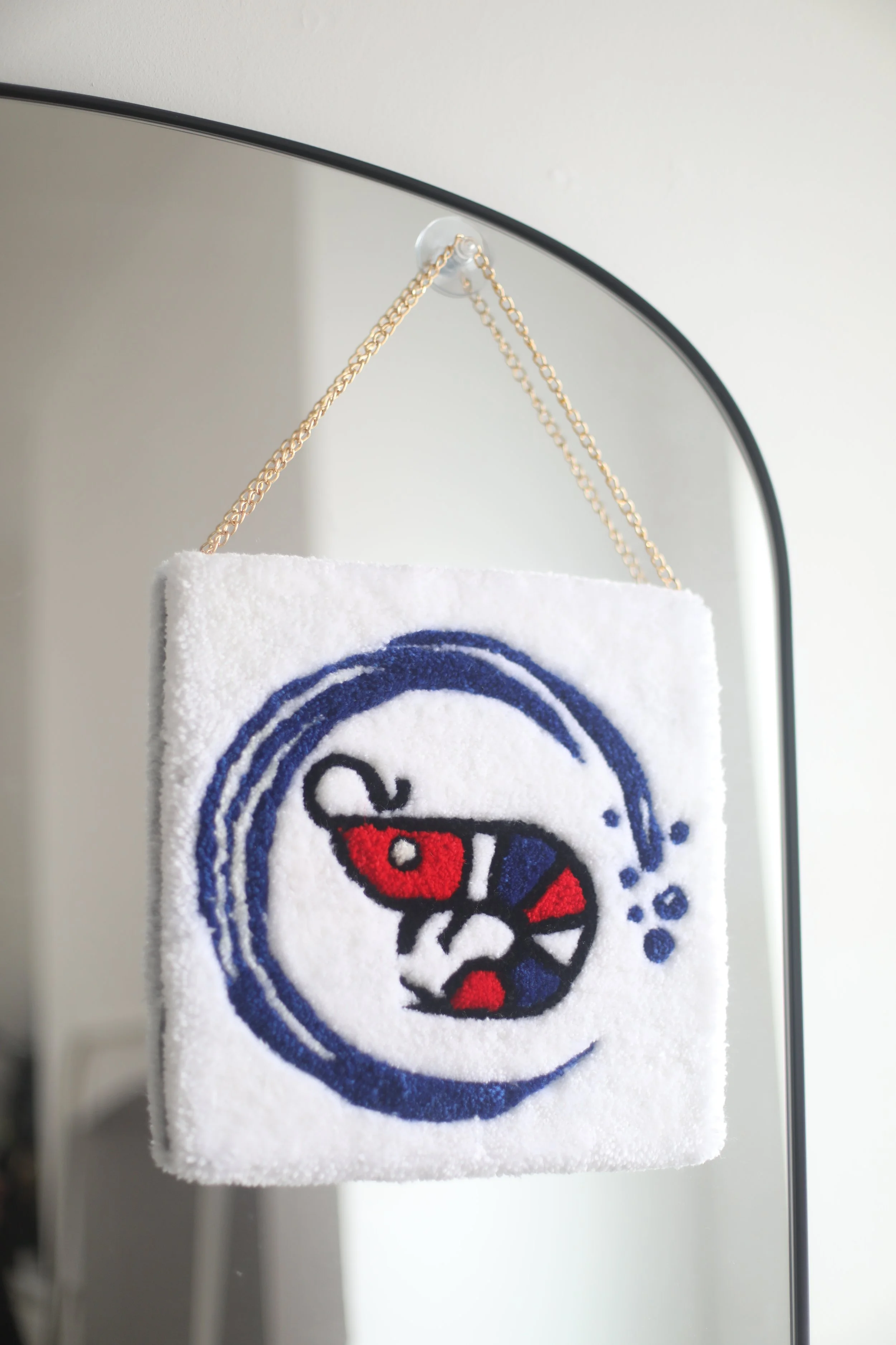A small wall hanging rug a prawn logo, hanging from a chain on a mirror stand, with a white wall in the background.