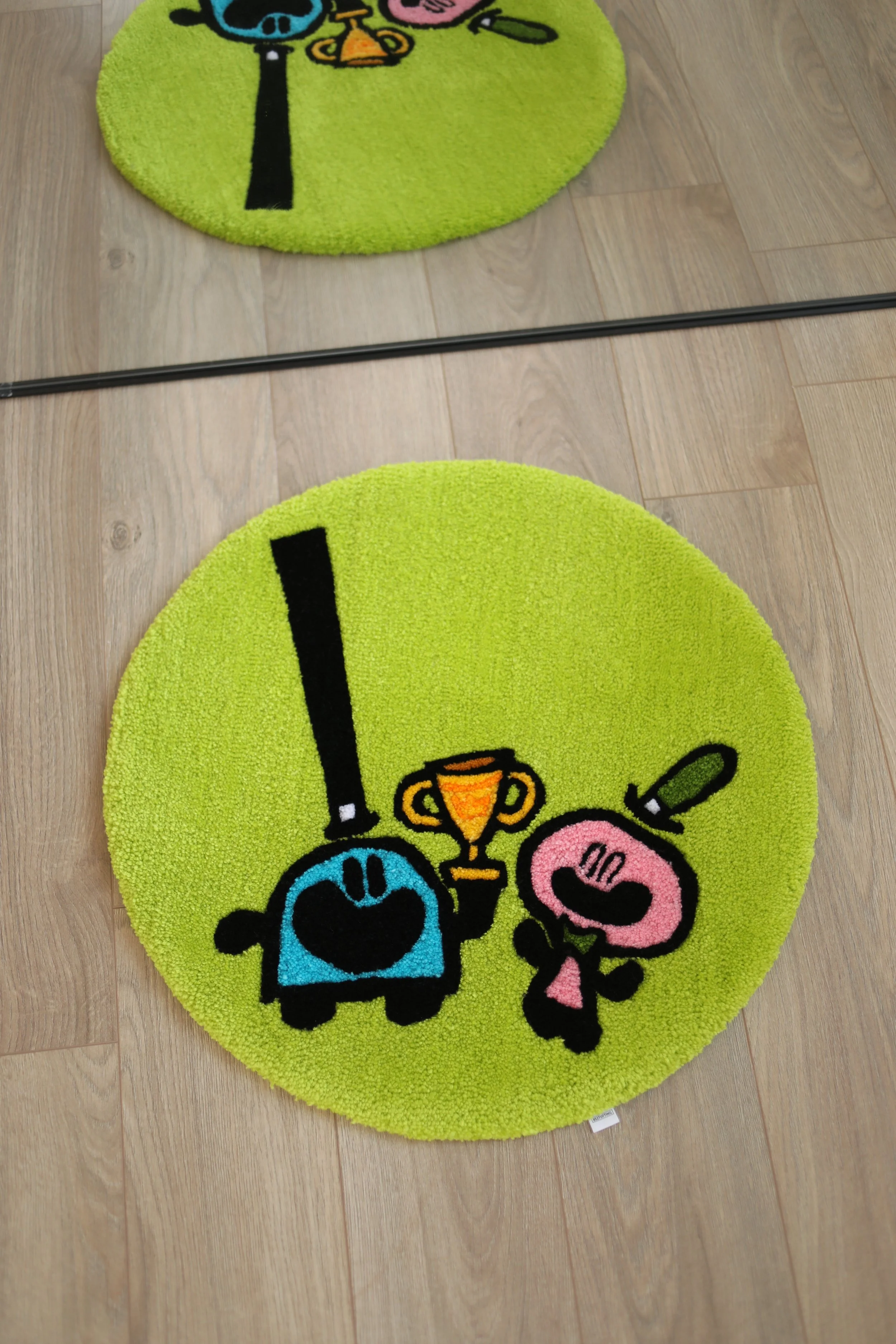 Bright green circular rug with cartoon characters, a chibi-style blue character with black hair and a pink character, each holding a trophy, on a wooden floor.