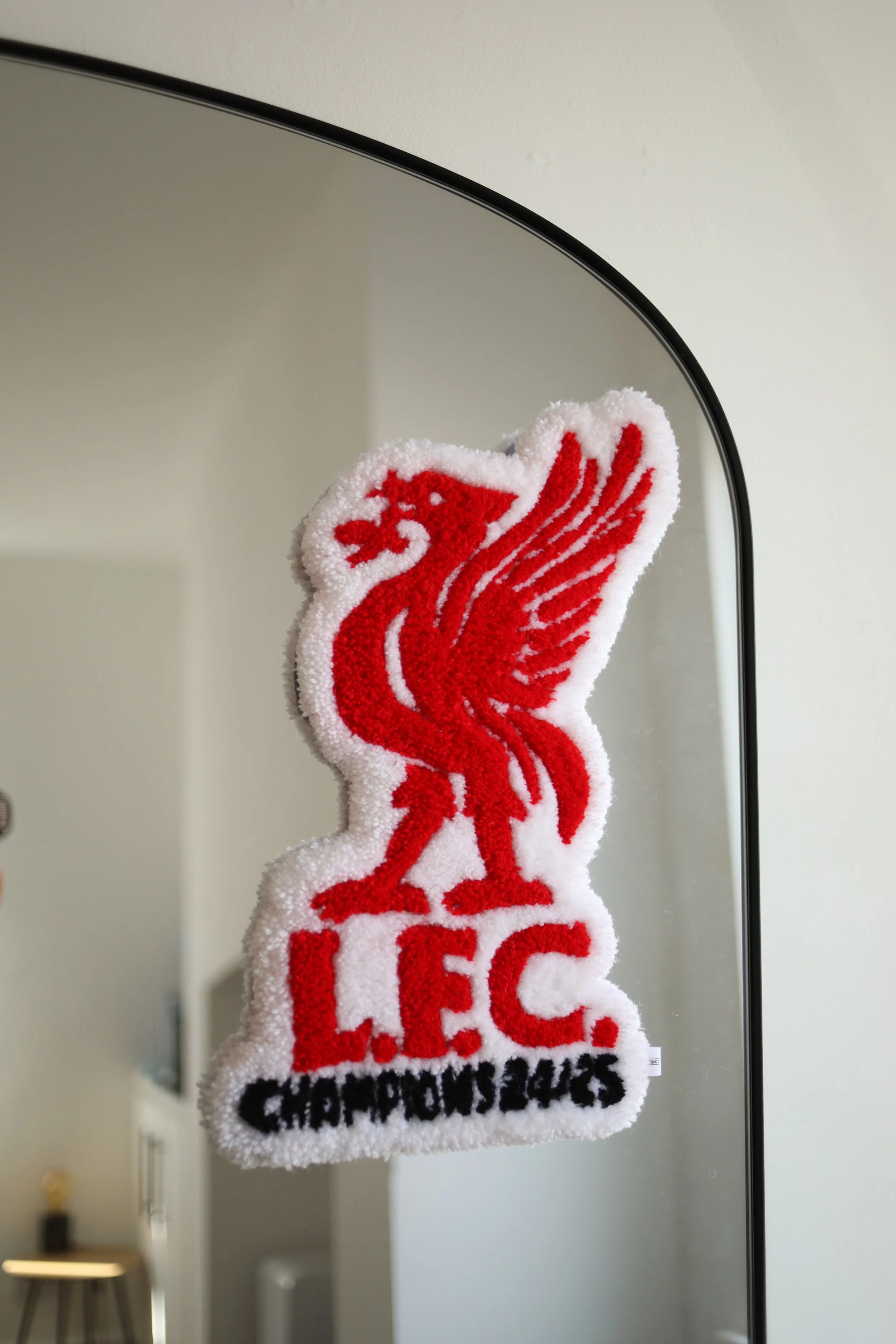 A fluffy towel with a red Liverpool Football Club logo and text, hanging in front of a mirror in a room with a side table and an artwork on the wall.