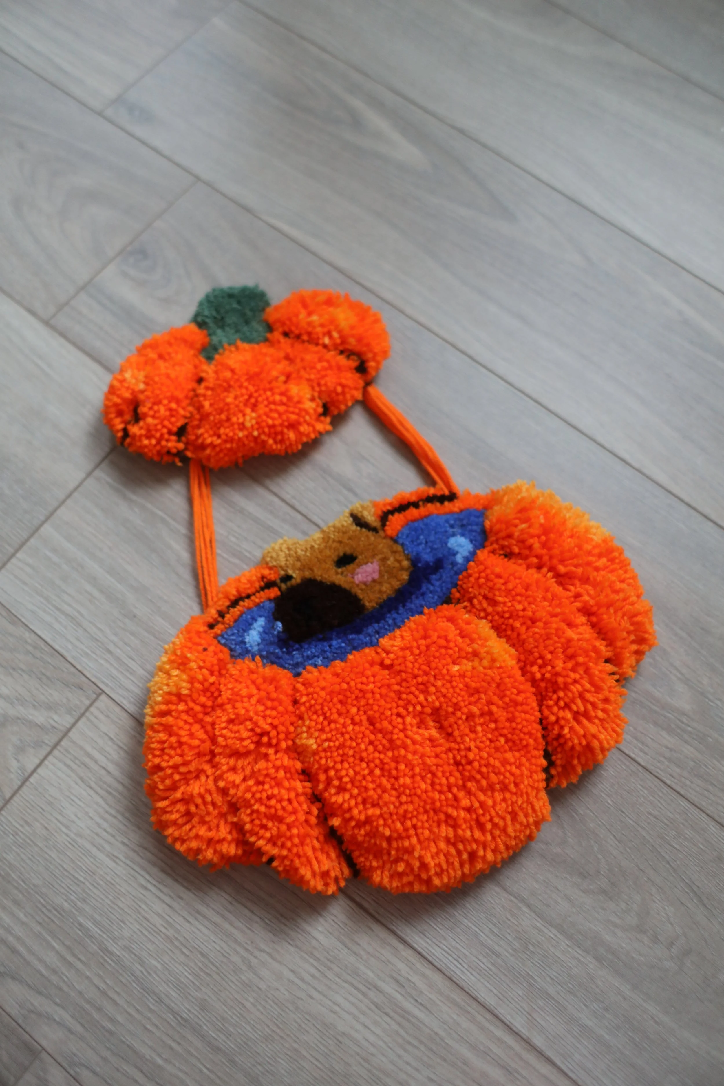 A plush toy shaped like a pumpkin with a smiling face, green stem, and orange body, lying on a wooden floor.