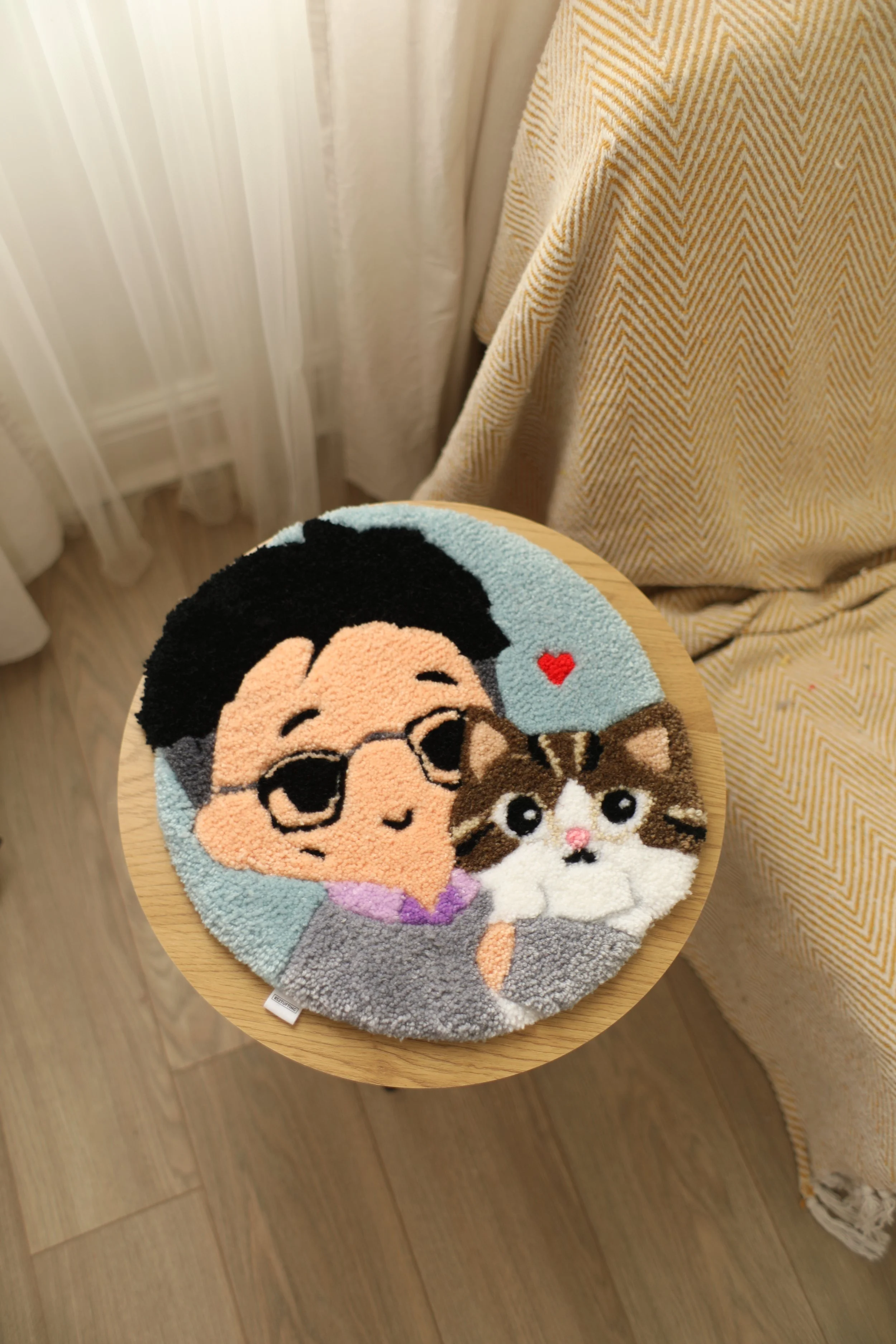 A round Rug with cartoon characters of a man and a cat, held in a person's lap, with a gold and cream zigzag patterned fabric on the right and a light-colored curtain in the background.