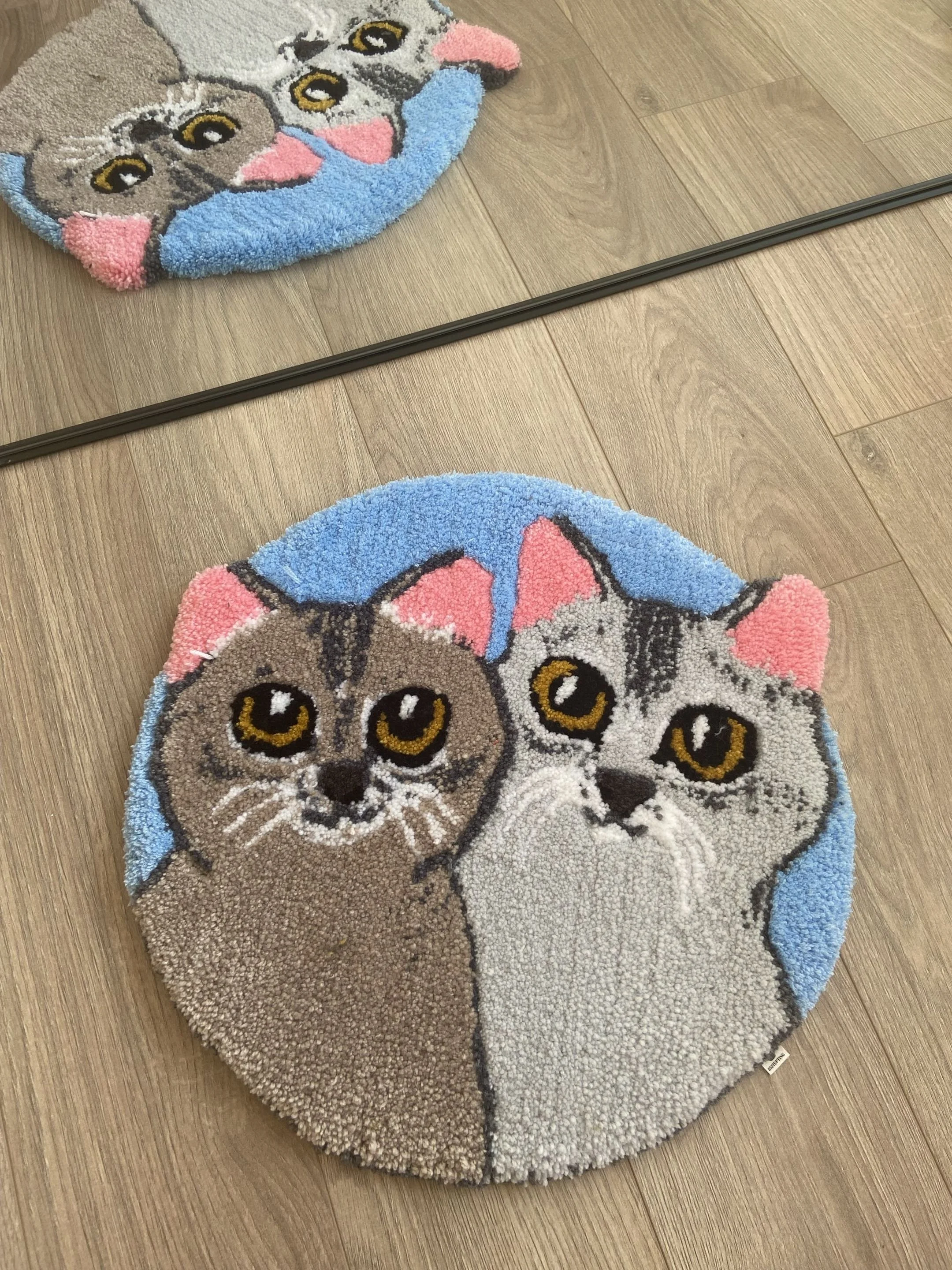 Round cat-themed rug with a kitten face, large eyes, pink nose, and blue and pink details, placed on a wooden floor.