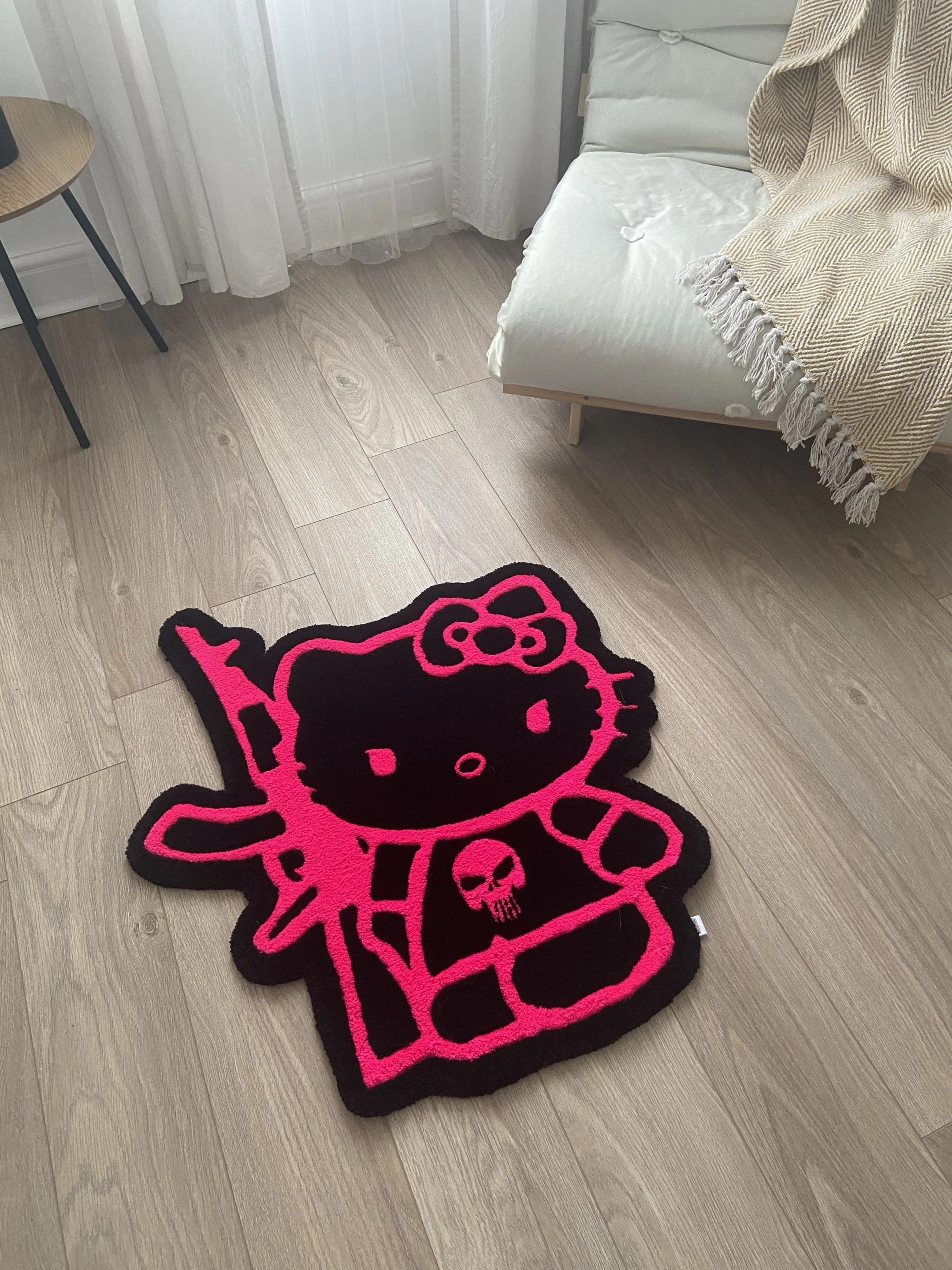 A pink and black Hello Kitty skeleton shaped rug on a light-colored wooden floor, with part of a beige and white blanket draped over a white bed and sheer white curtains in the background.